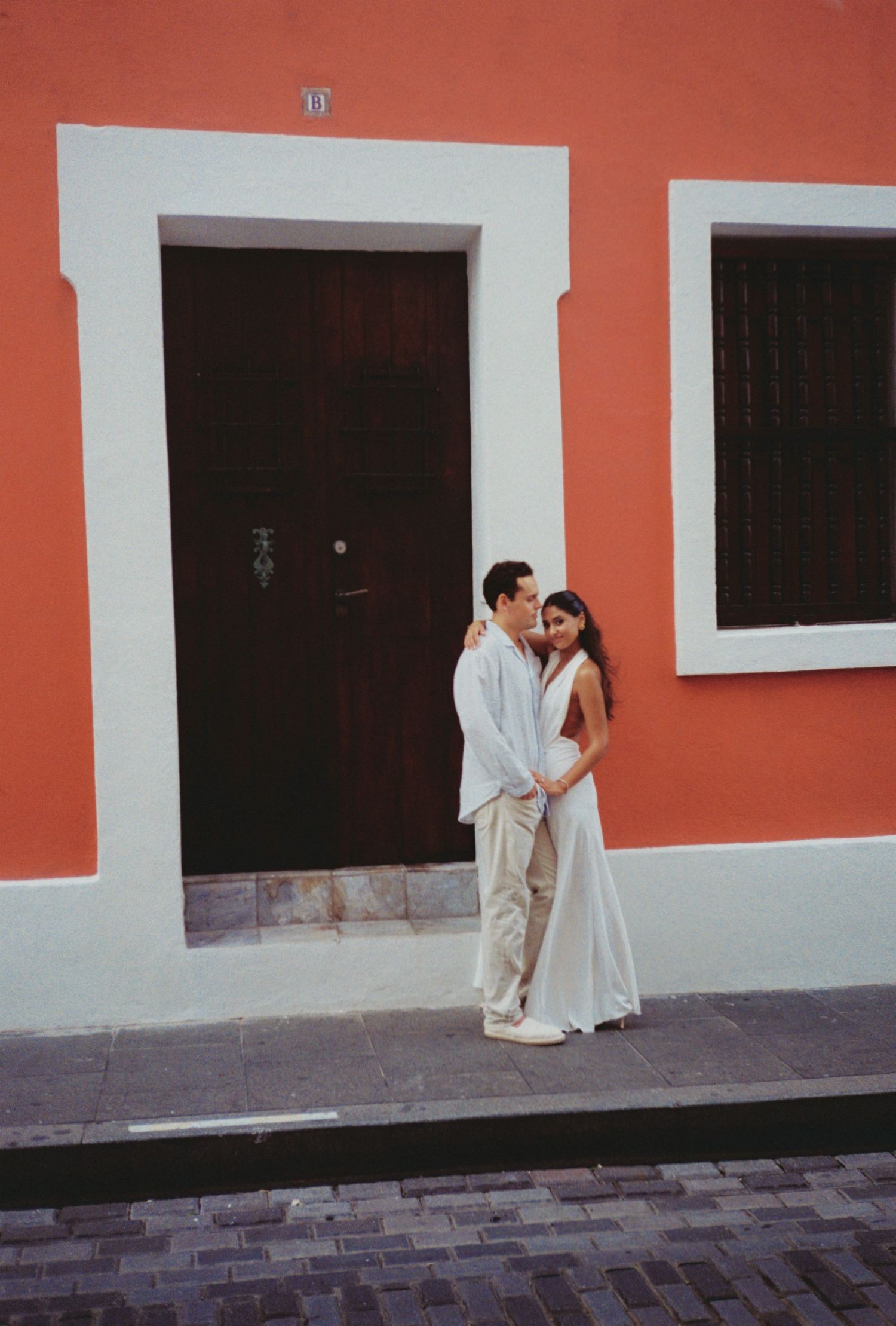 Couple in wedding attire embracing outside a colorful coral building with a dark wooden door in Old San Juan.
