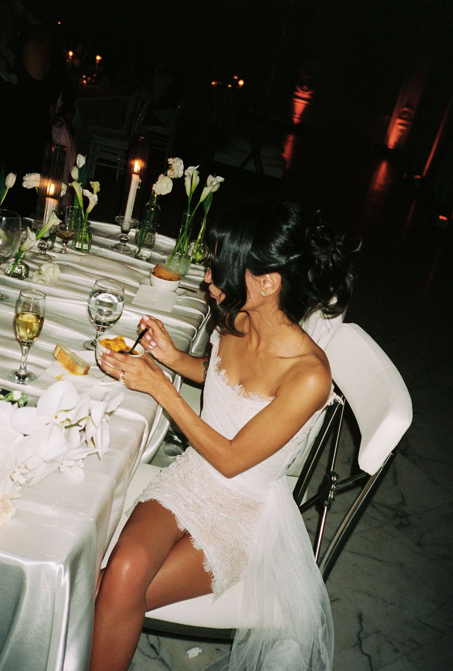 Bride in white lace dress sitting at elegant candlelit wedding reception table with champagne and floral centerpieces.