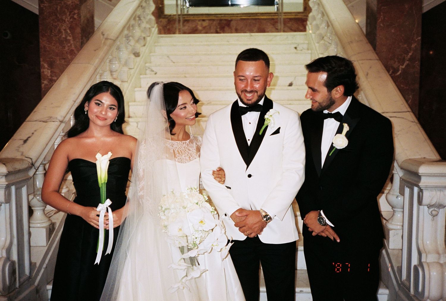Wedding party posing on grand staircase, bride in white gown with veil, groom in white tuxedo, guests in formal black attire.