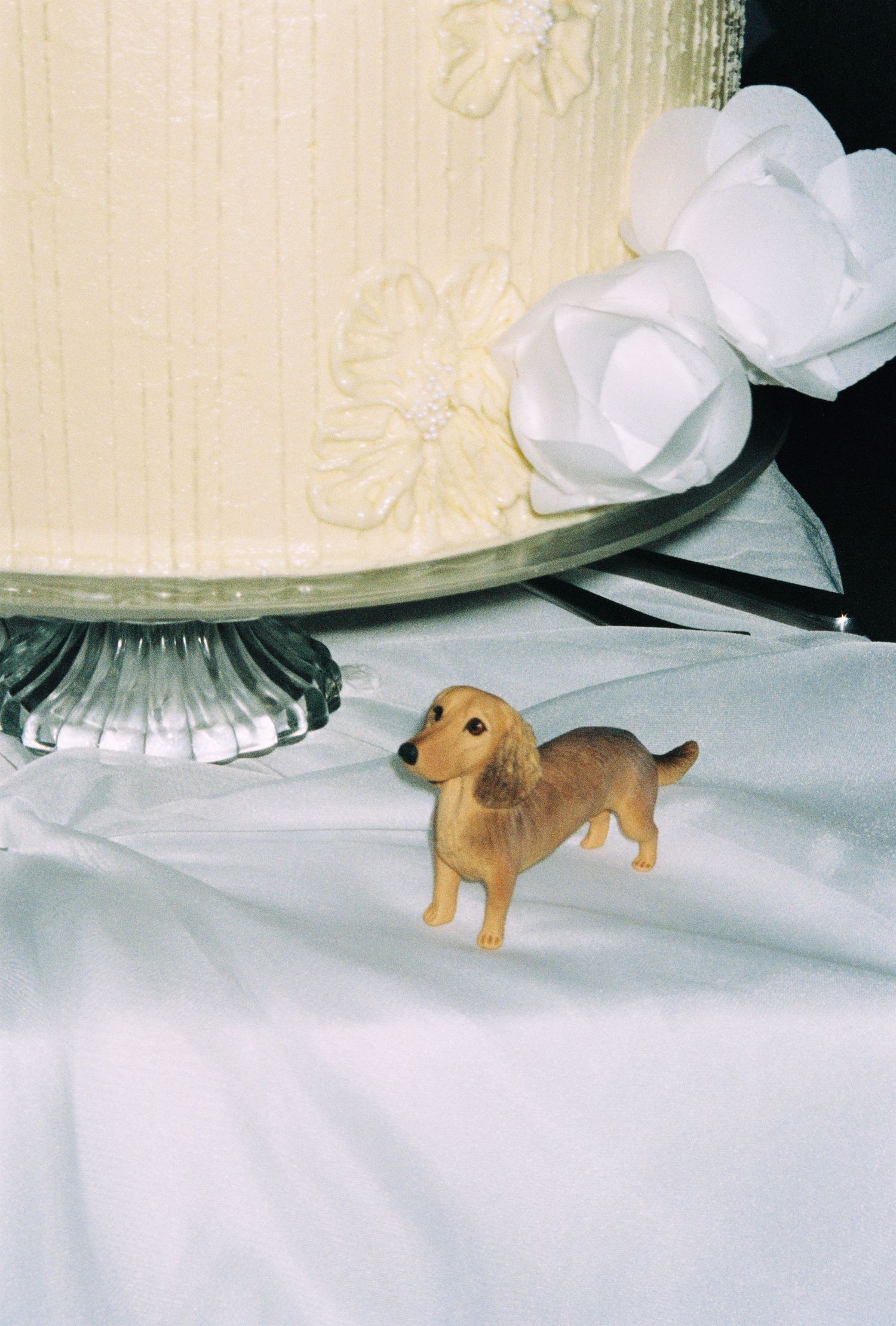 A small dachshund figurine stands beside an elegant white wedding cake on a glass pedestal with floral decorations.