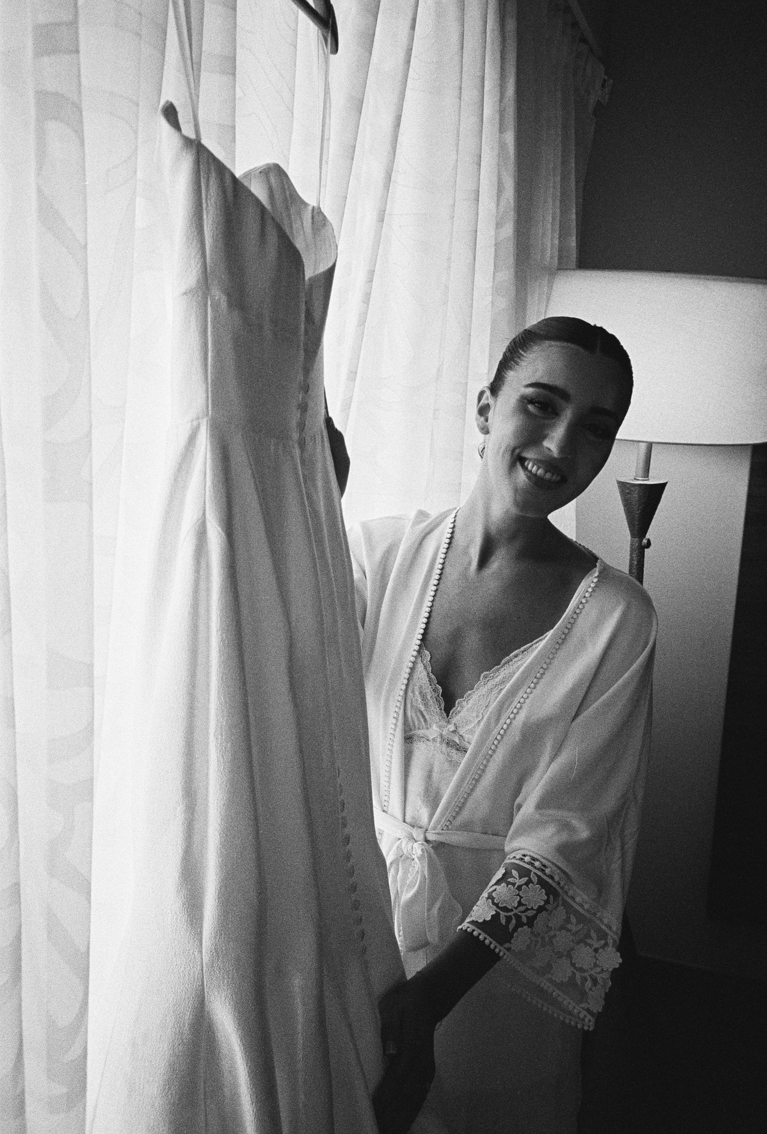 Smiling bride in white robe standing beside her wedding dress hanging by a curtained window, black and white photo.
