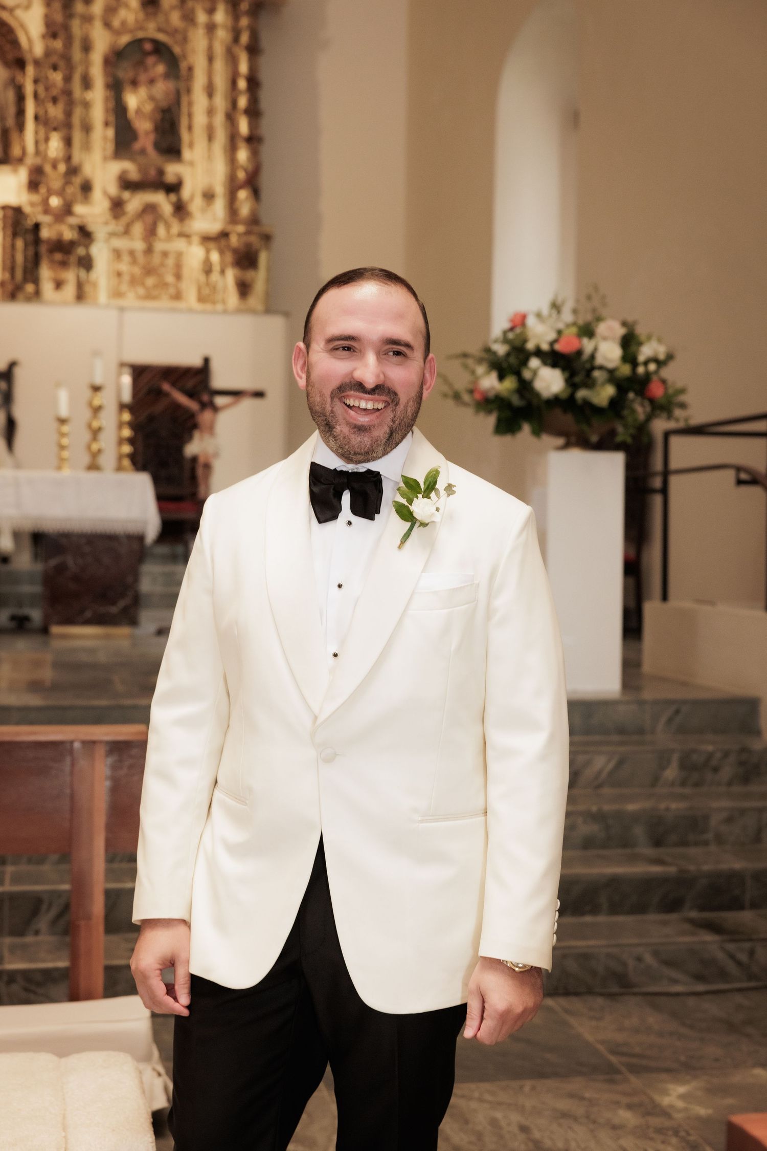 A groom in a white tuxedo jacket and black bow tie stands in an elegant church setting.