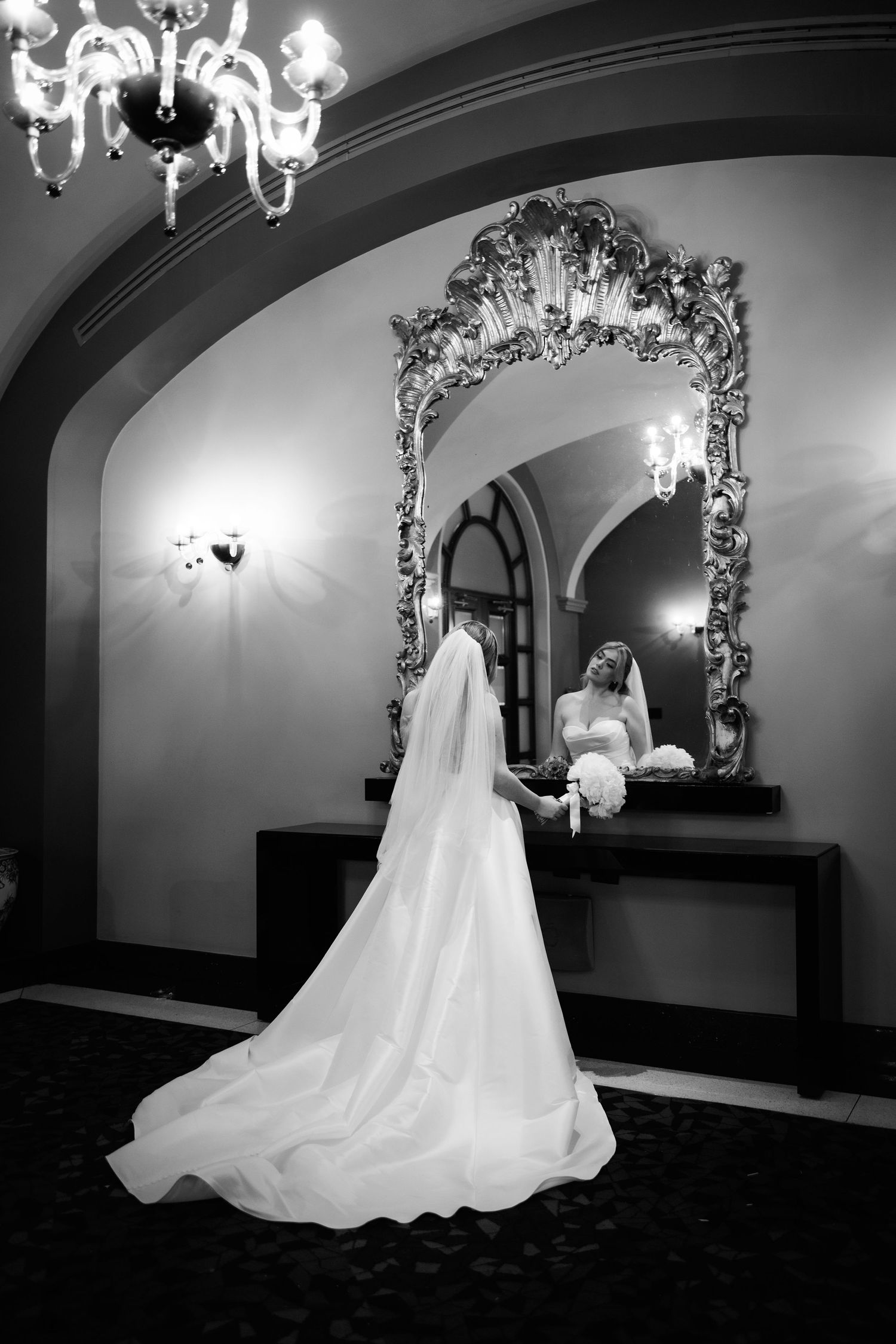 Black and white photo of bride in flowing gown looking at ornate mirror under chandelier lighting.
