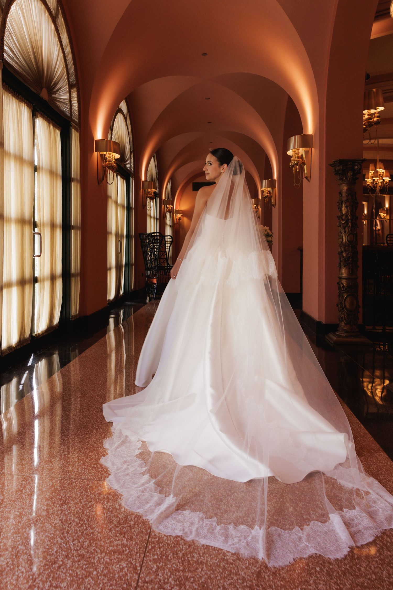A bride in a flowing white wedding gown walks down an elegant arched hallway with pink walls and polished marble floors.