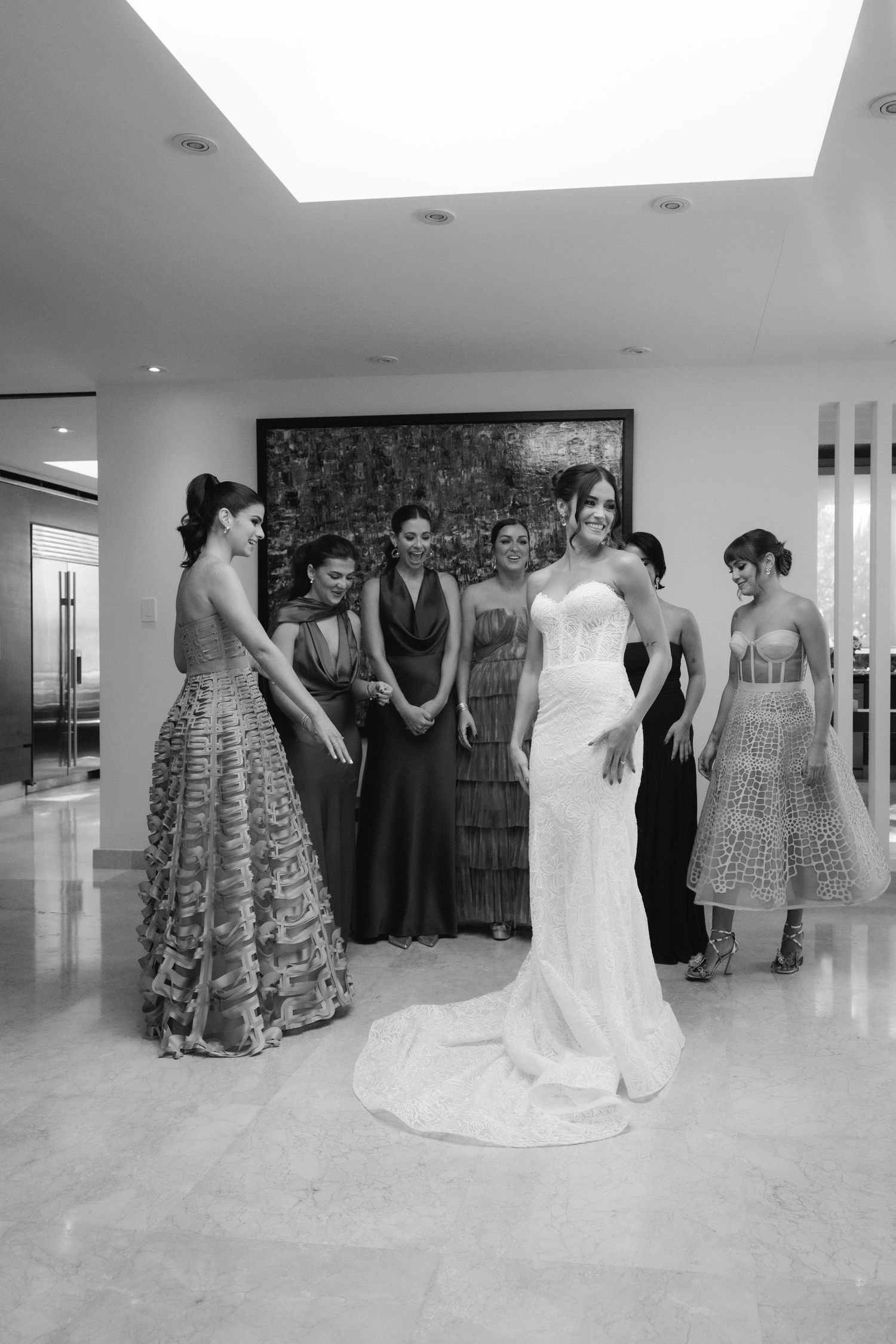 Wedding party members in colorful dresses gather around bride in spacious white room.