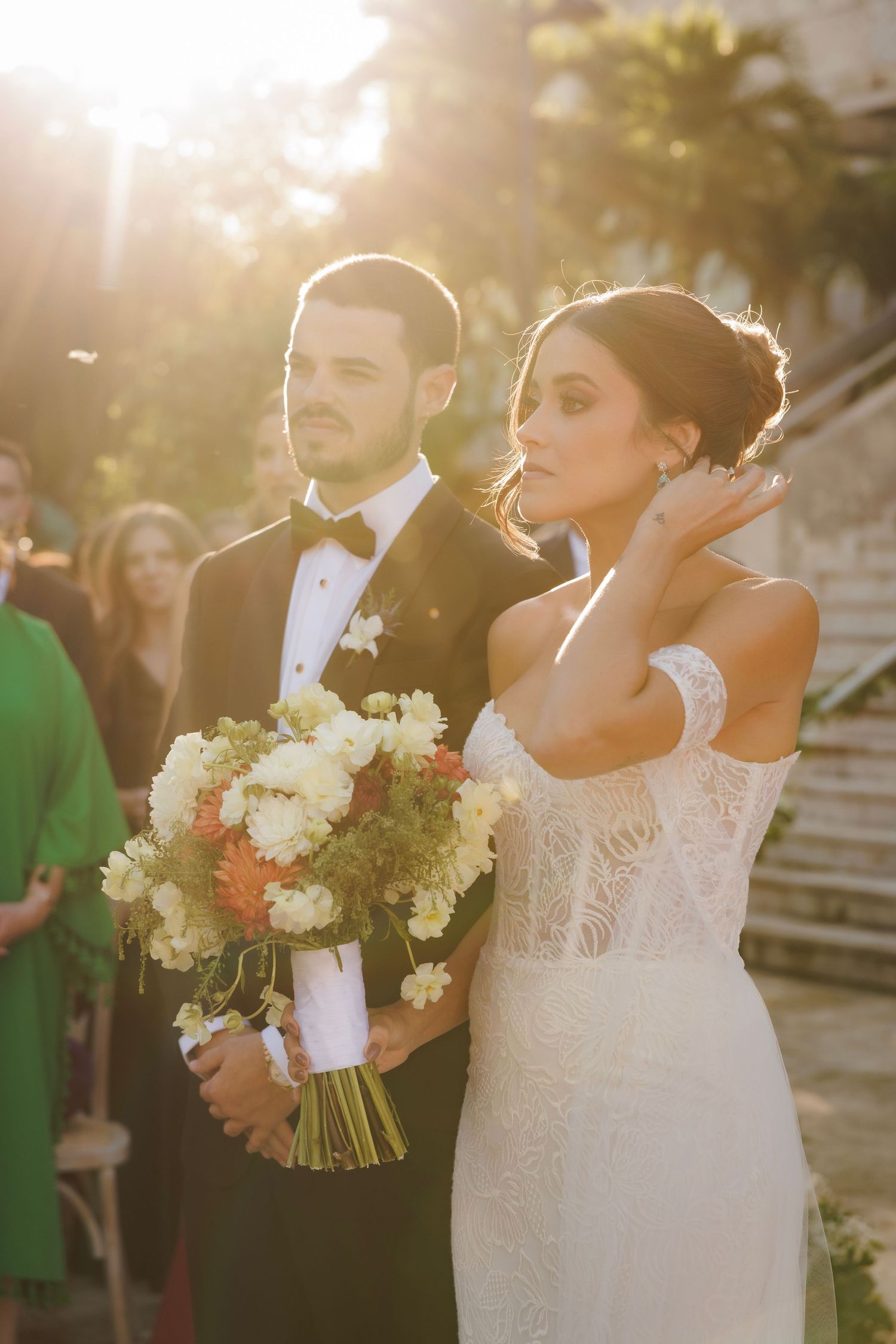 Wedding couple in formal attire holding white floral bouquet during golden hour.
