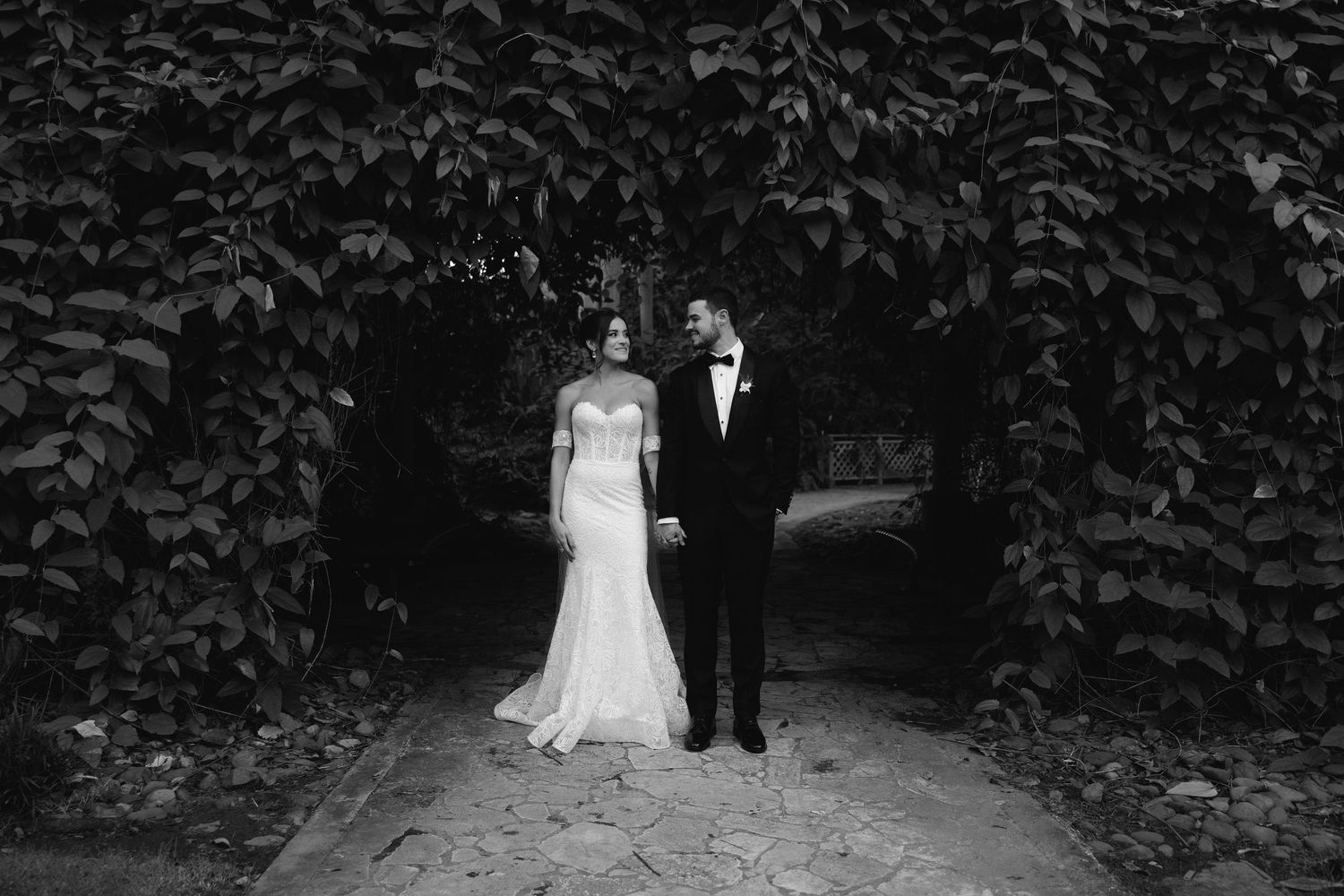 A black and white wedding photo series showing a couple walking through a lush garden pathway in formal attire.