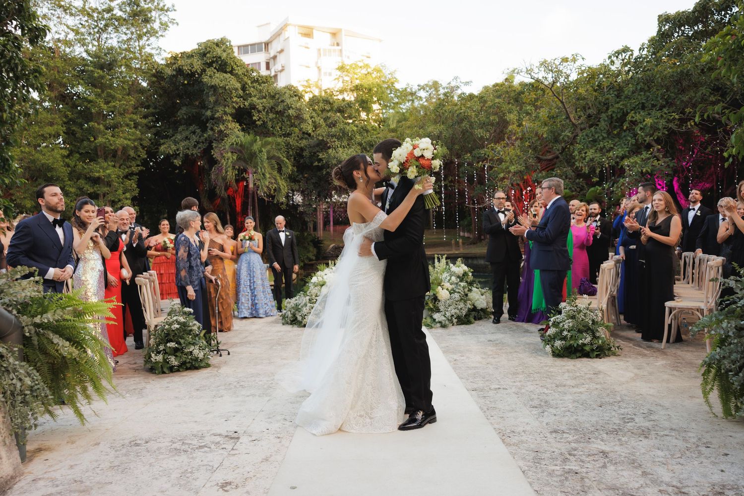 Wide shot of wedding ceremony with guests watching couple from garden pathways.