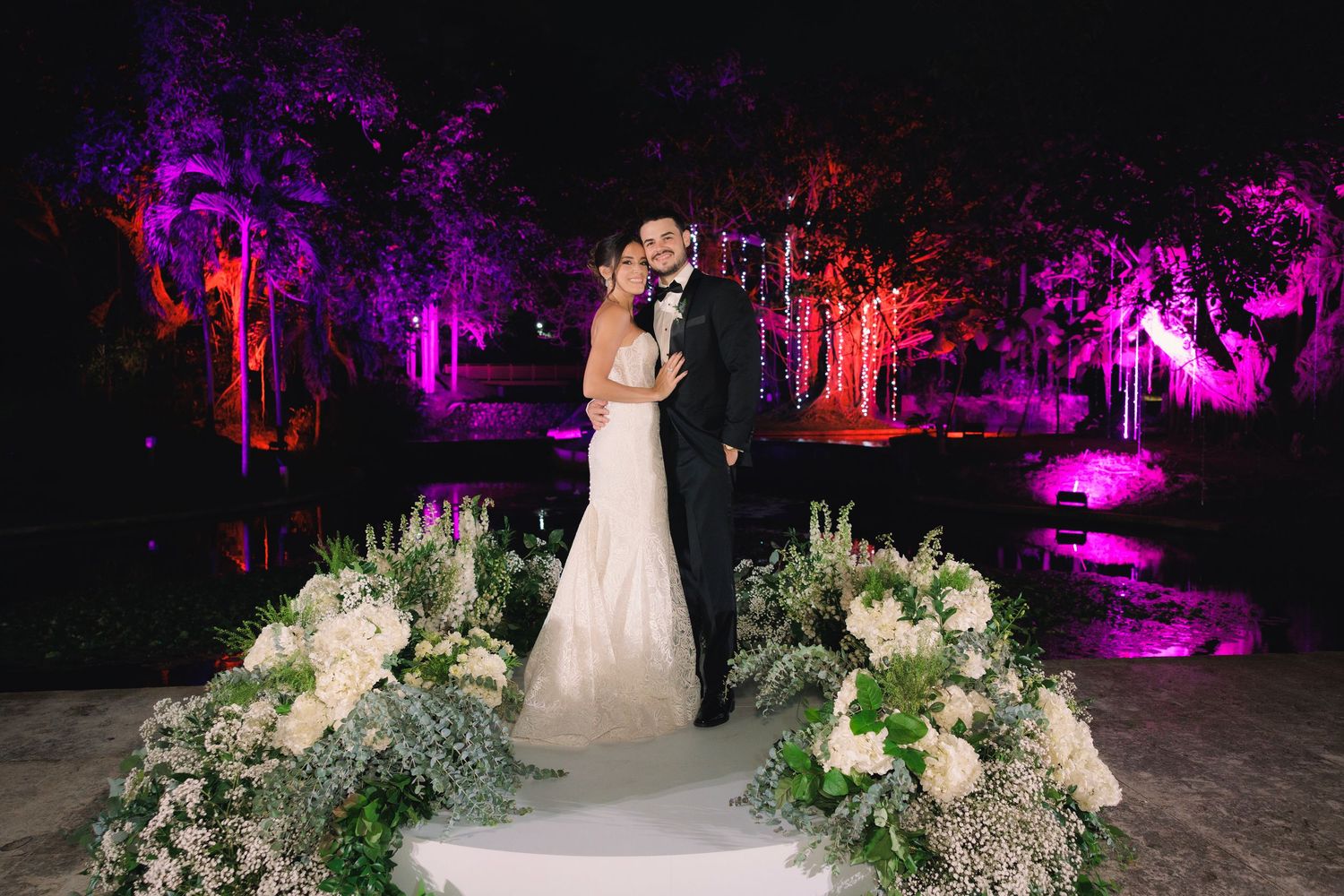 Outdoor wedding scene with elegant floral arrangements illuminated by dramatic purple and red lighting.