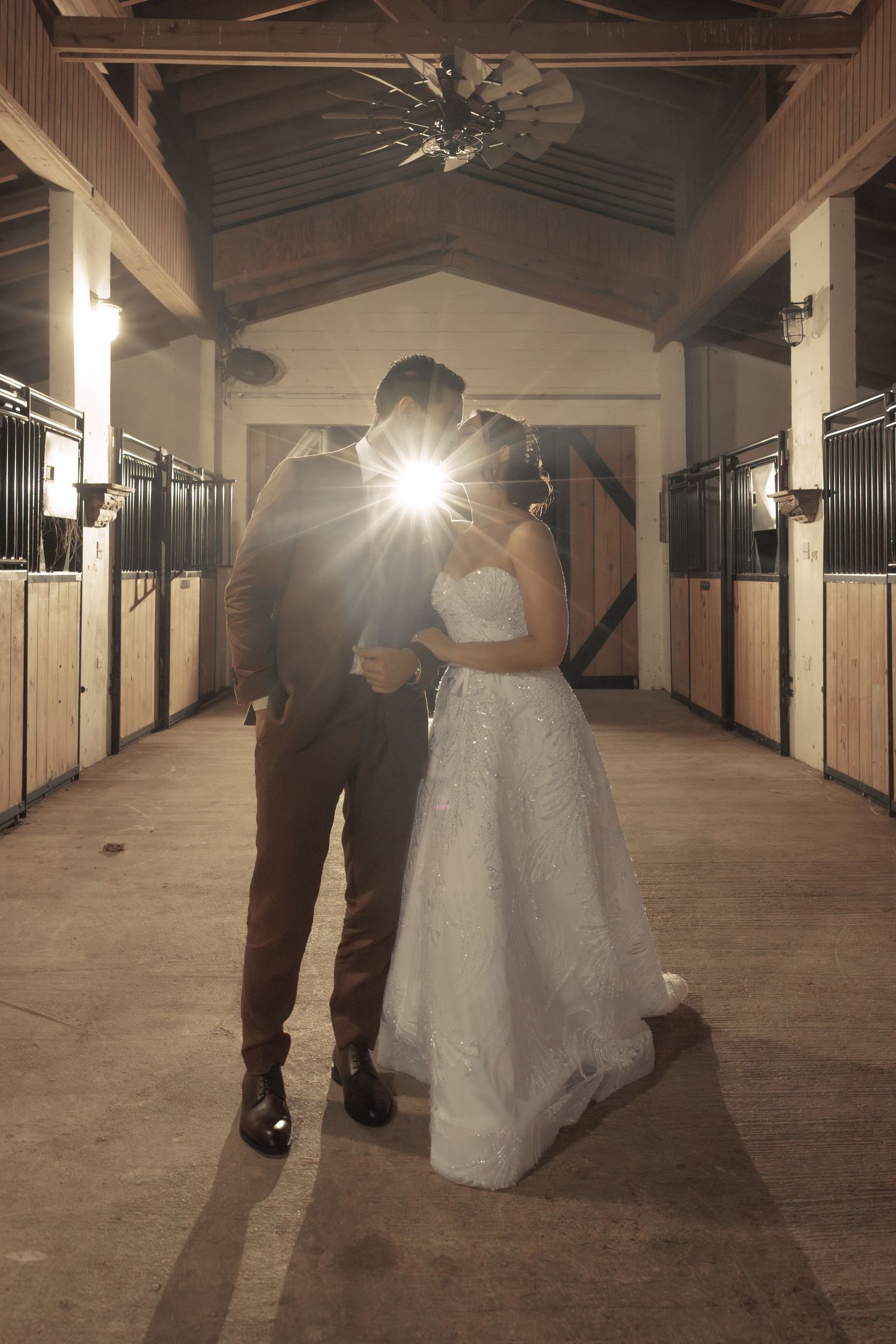 Wedding couple dances in sunlit rustic barn hallway with wooden stalls.