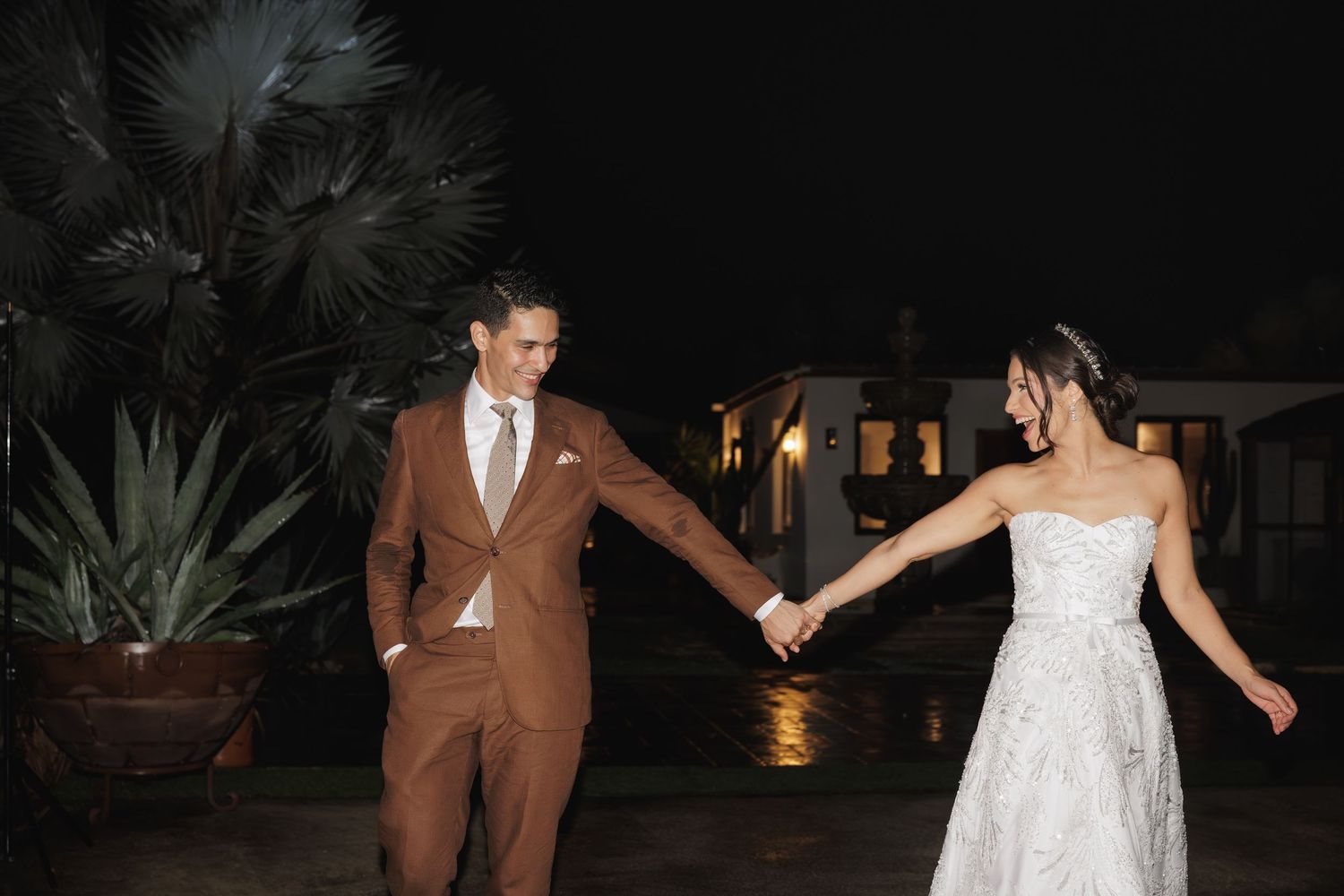 A couple dances together at an outdoor evening wedding reception surrounded by tropical plants and soft lighting.