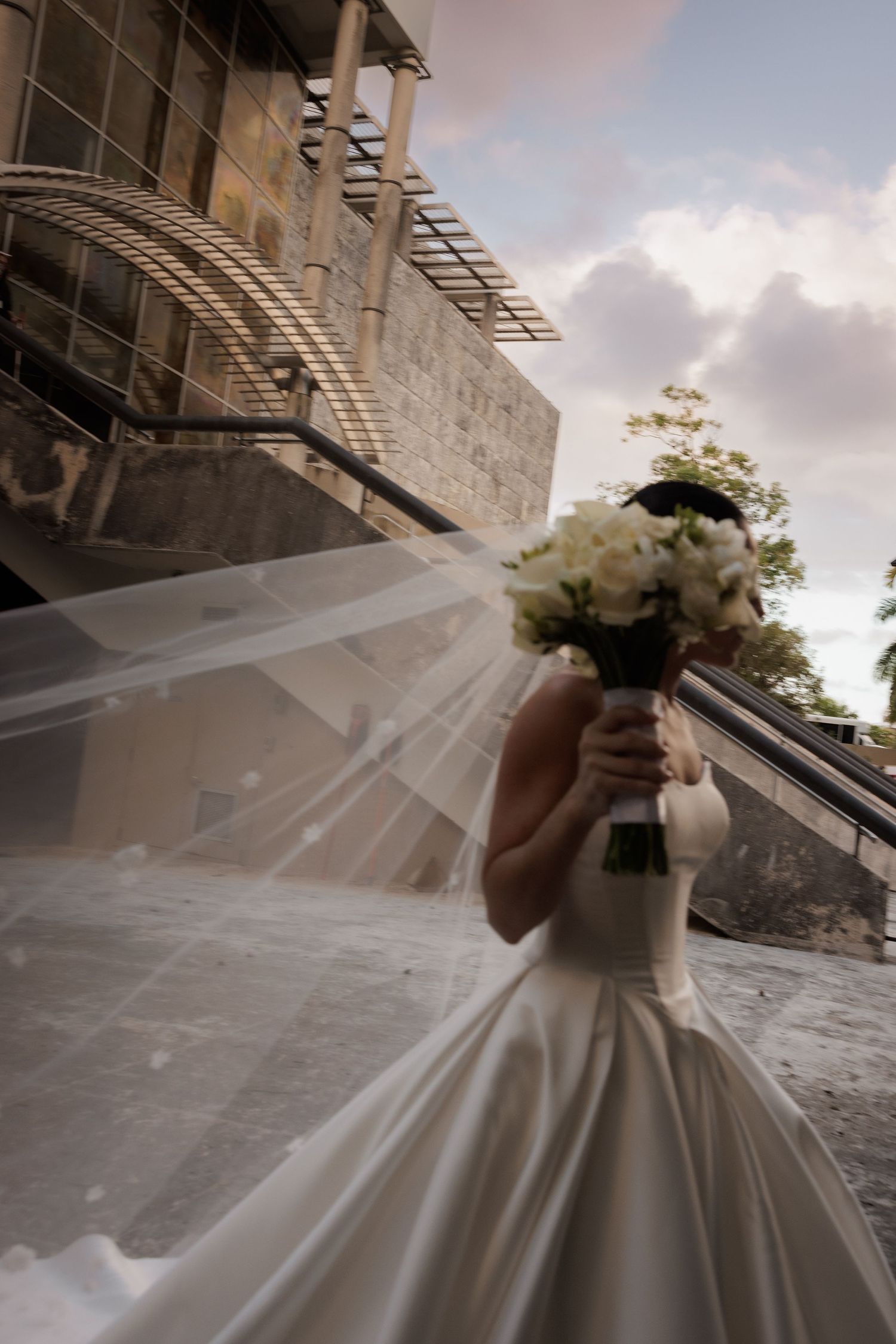 Bride in flowing white gown holding bouquet against stone building exterior.