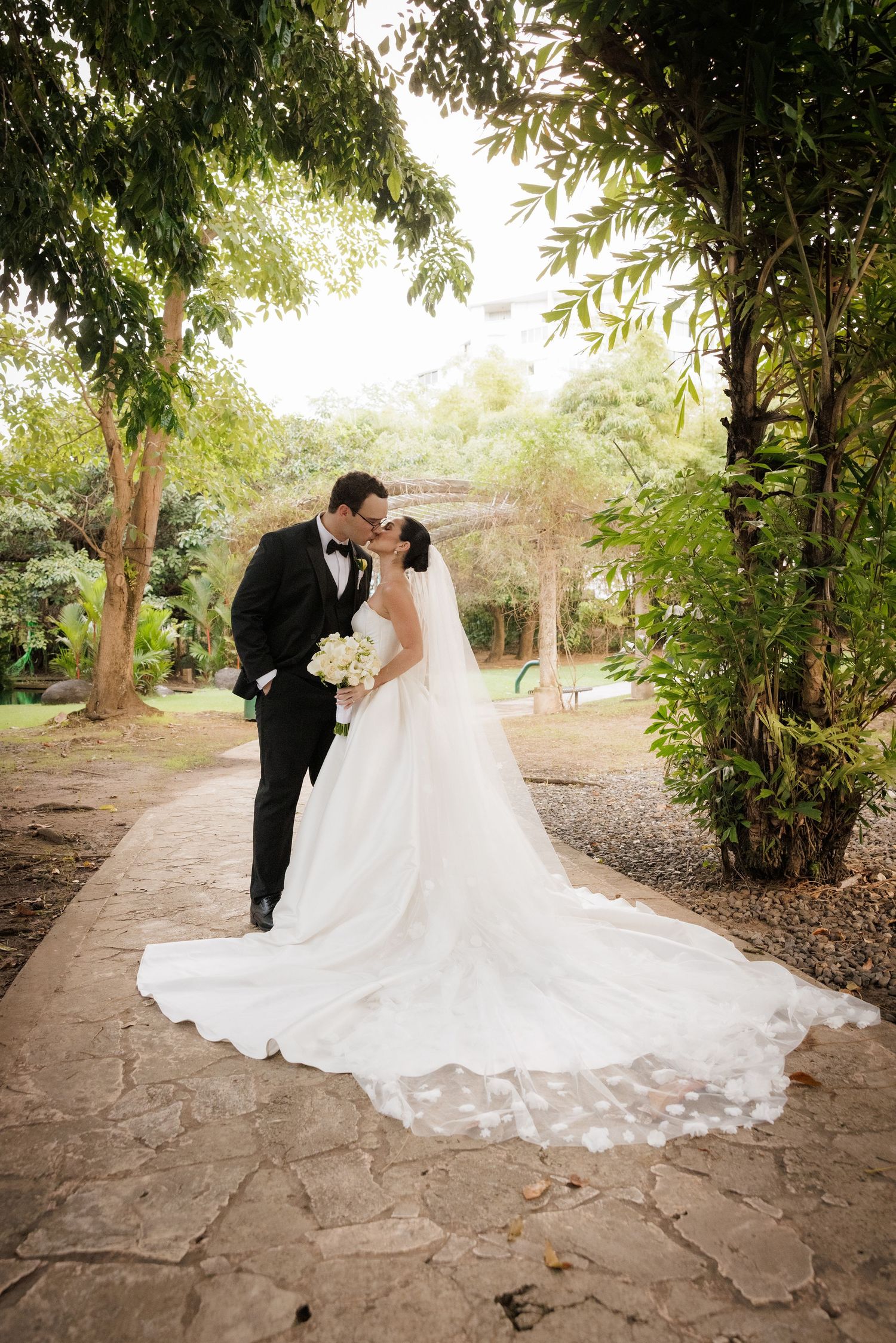 A romantic wedding couple shares intimate moments in a lush garden setting with stone pathways and greenery.