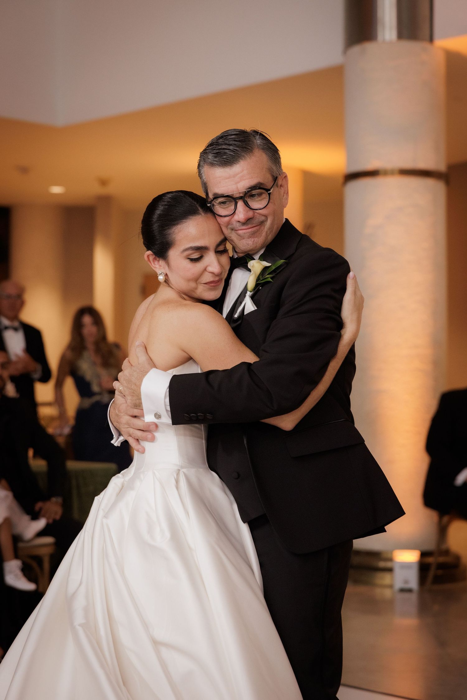 Newlyweds embrace during their first dance in an elegant venue with white pillars and warm lighting.