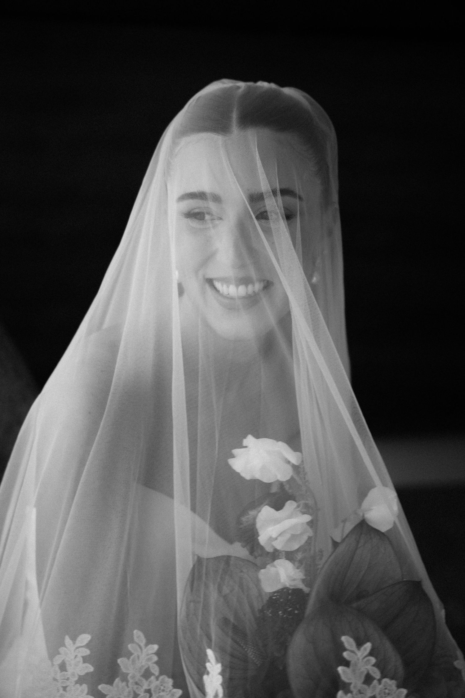 Black and white portrait of a smiling bride wearing a sheer veil over her face, holding a bouquet of roses.