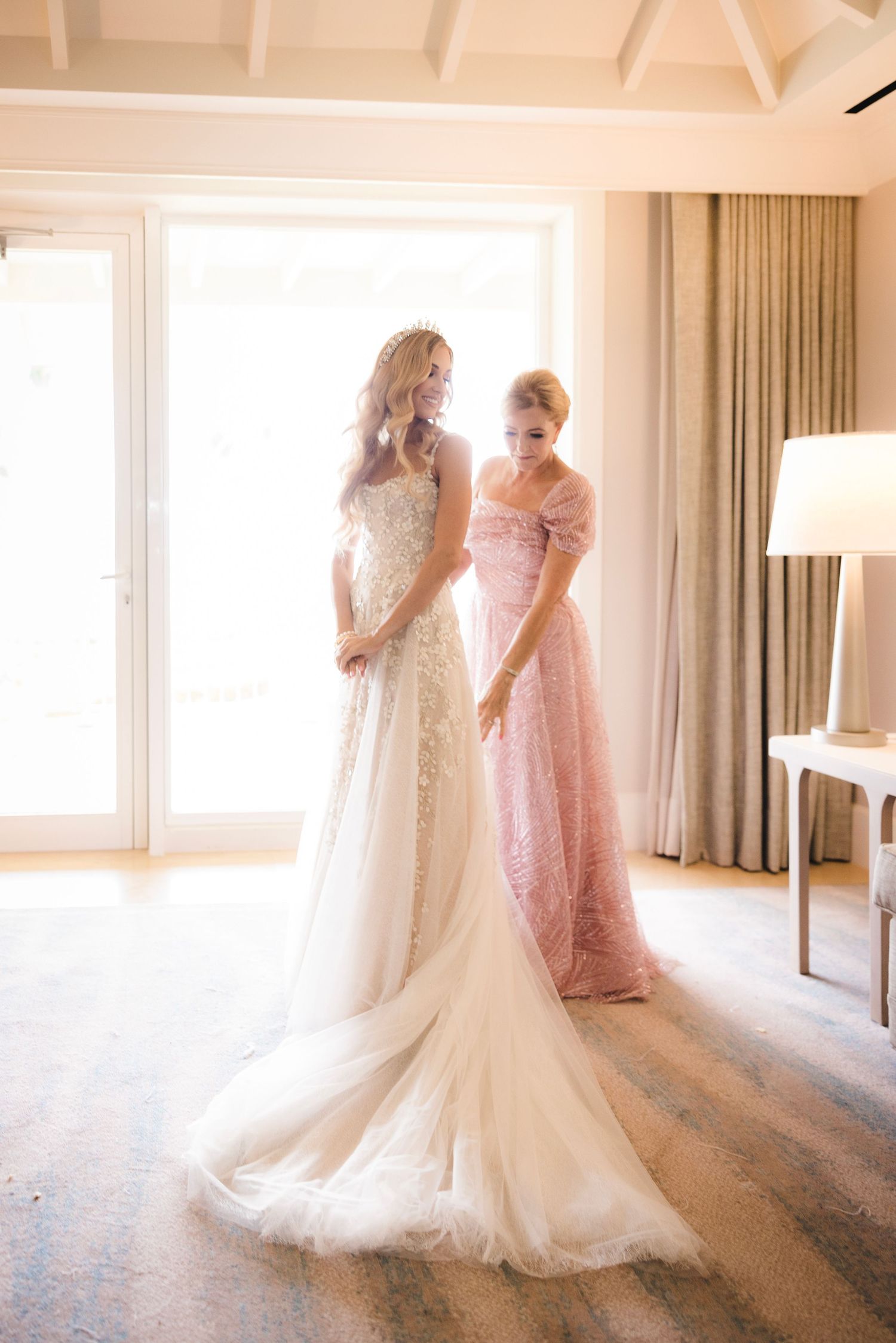 Two figures in elegant gowns, one in a flowing white wedding dress and one in pink lace, stand together in sunlit room.