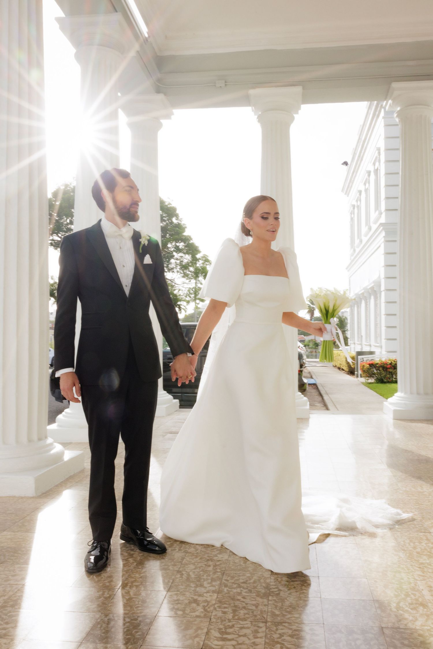 A couple in wedding attire walks along a sunlit colonnade with bright white pillars.