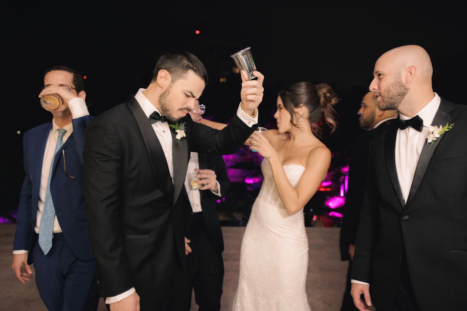 Wedding guests in formal attire toasting and celebrating at an evening reception with purple accent lighting.