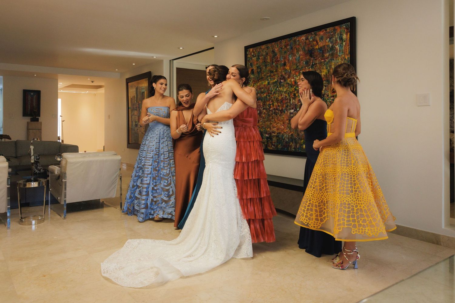 Bride in white lace gown celebrates with bridesmaids wearing colorful dresses during wedding preparations.