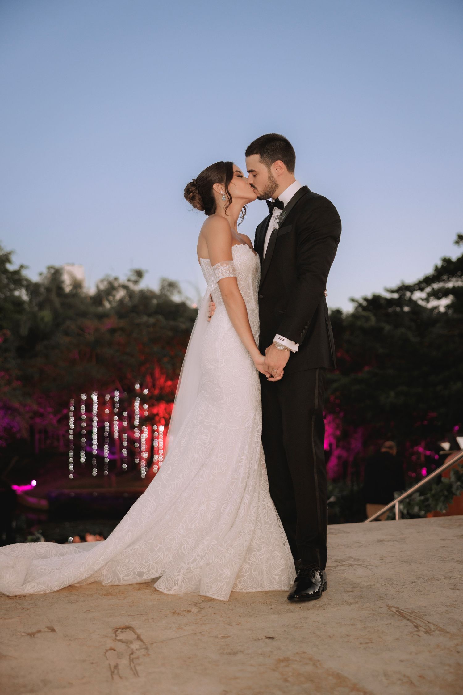 A romantic wedding couple shares a kiss at dusk with purple lighting in the background and a flowing white gown.