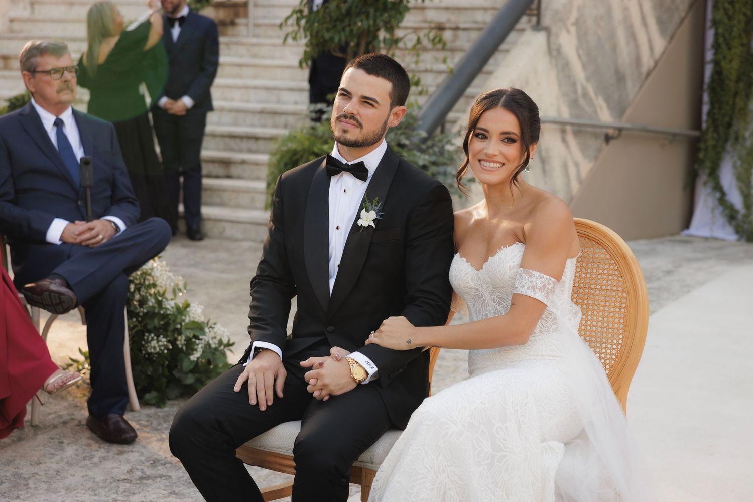 A bride in a strapless white gown and groom in black tuxedo sit together during an outdoor wedding ceremony.