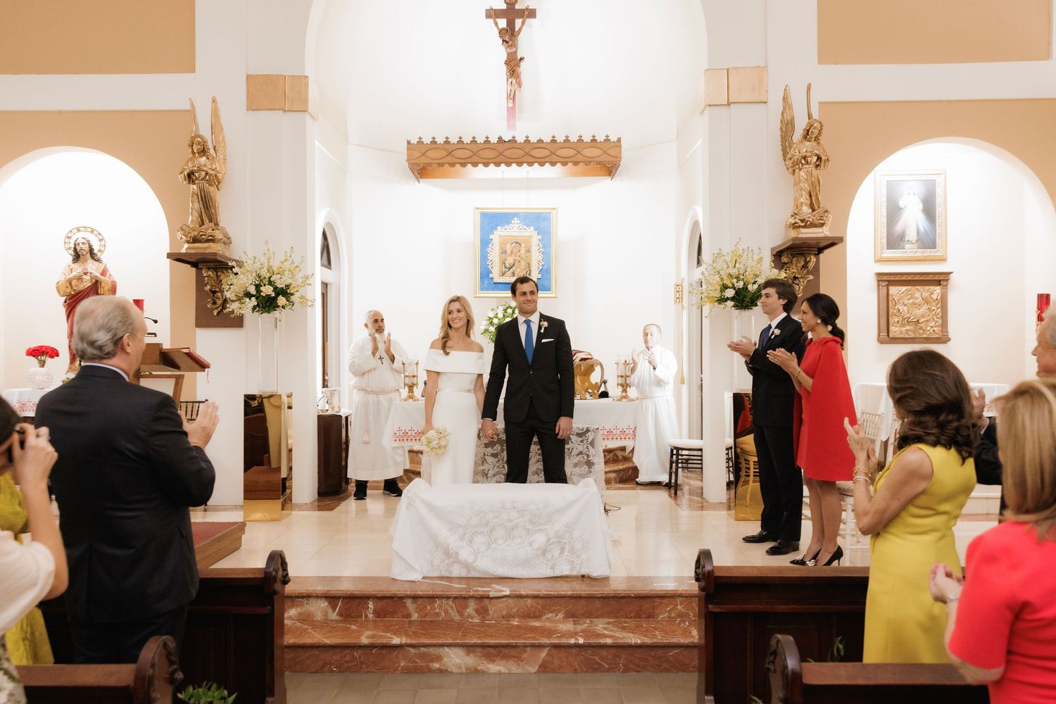 A wedding ceremony takes place inside a traditional Catholic church with ornate architecture and floral decorations.