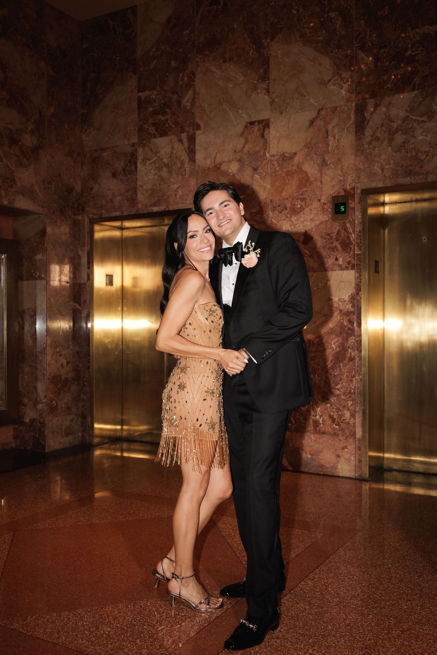 A couple poses together by gold elevator doors in an elegant marble-walled venue.