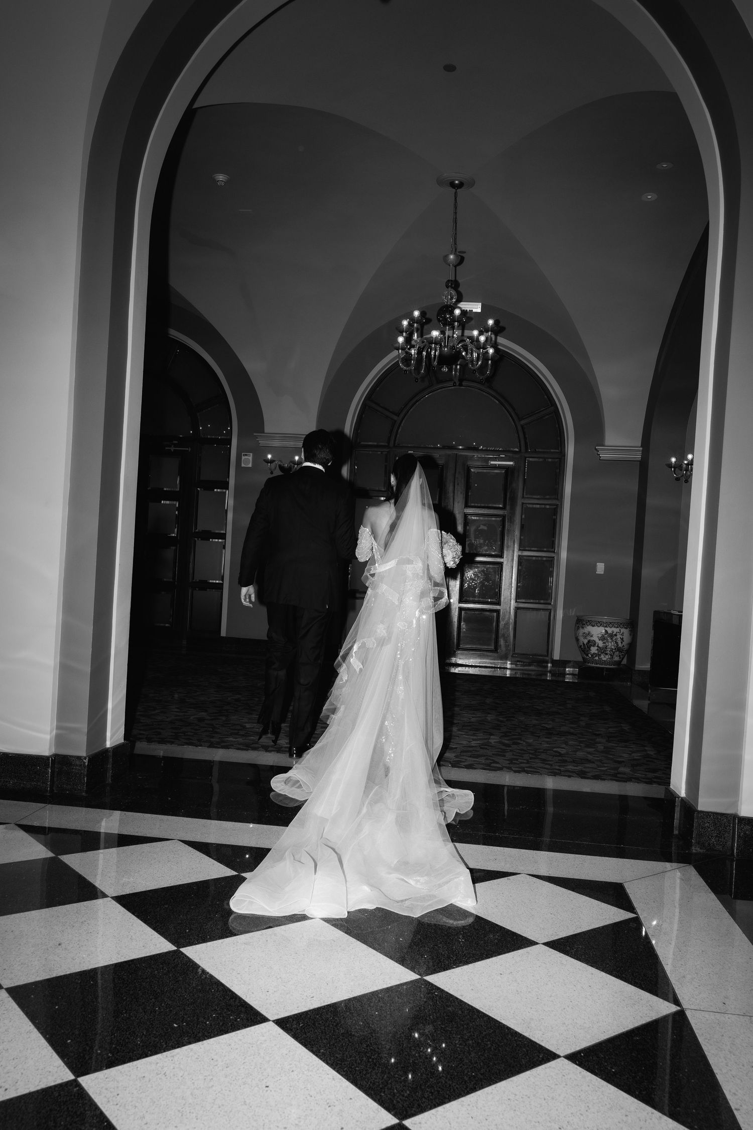 A couple in wedding attire walk across a checkered floor beneath an elegant arched hallway with a chandelier.