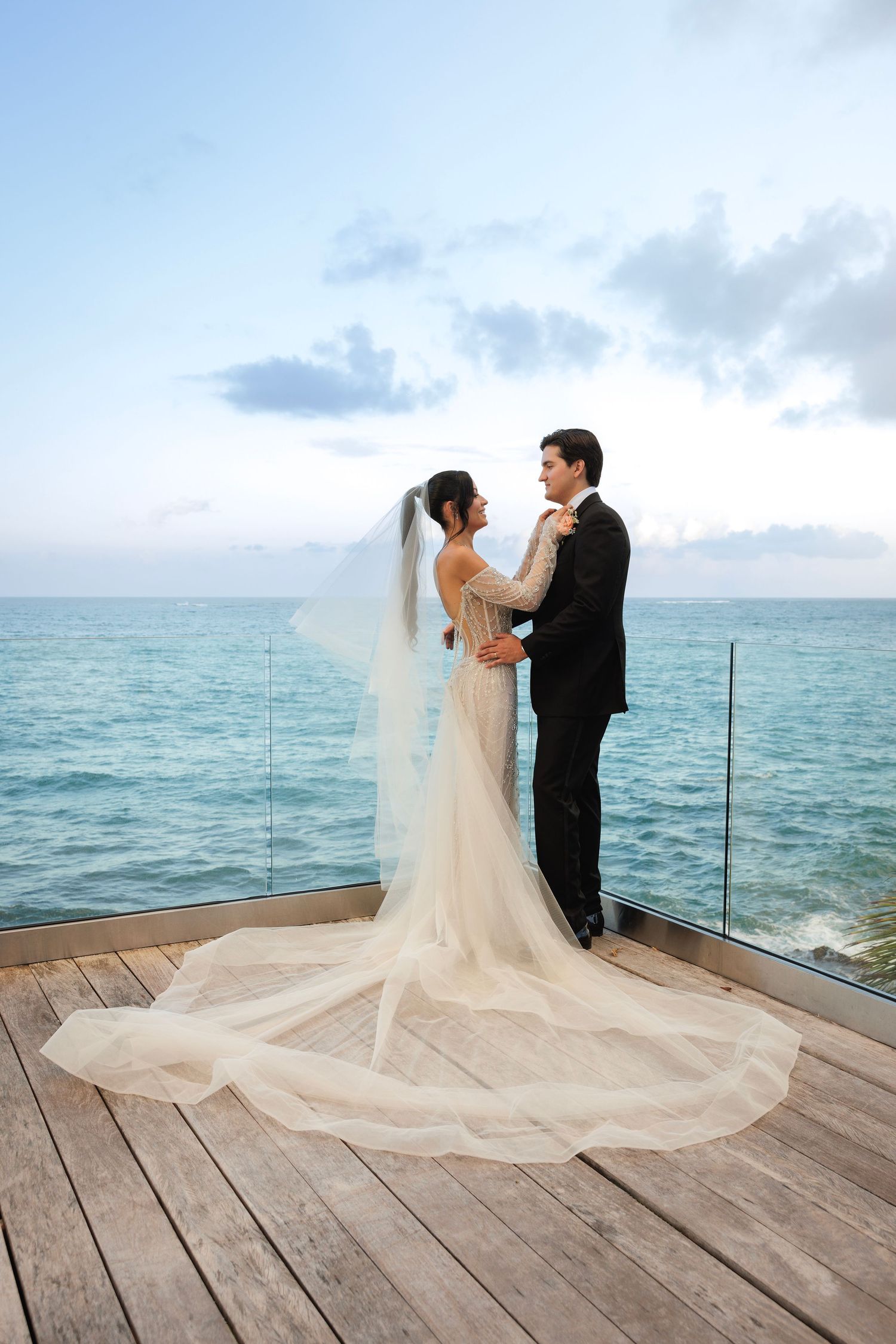 A couple dances on a wooden deck overlooking the ocean during their wedding, with the bride's veil trailing elegantly behind.