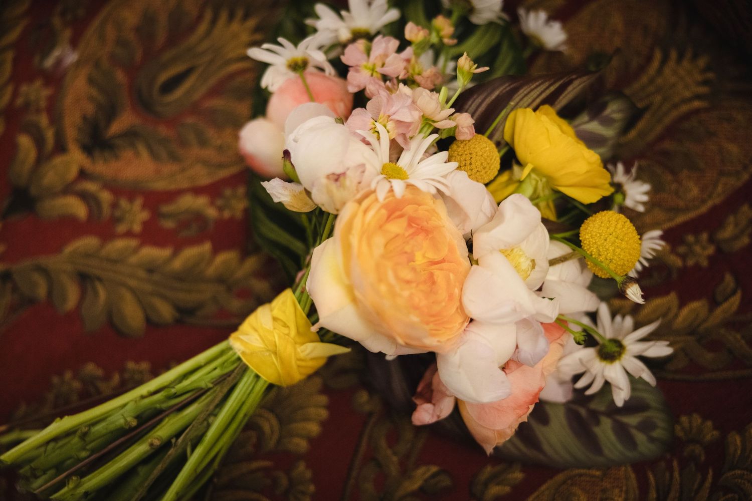 A romantic bouquet featuring coral roses, white daisies, and yellow ranunculus flowers against a dark background.
