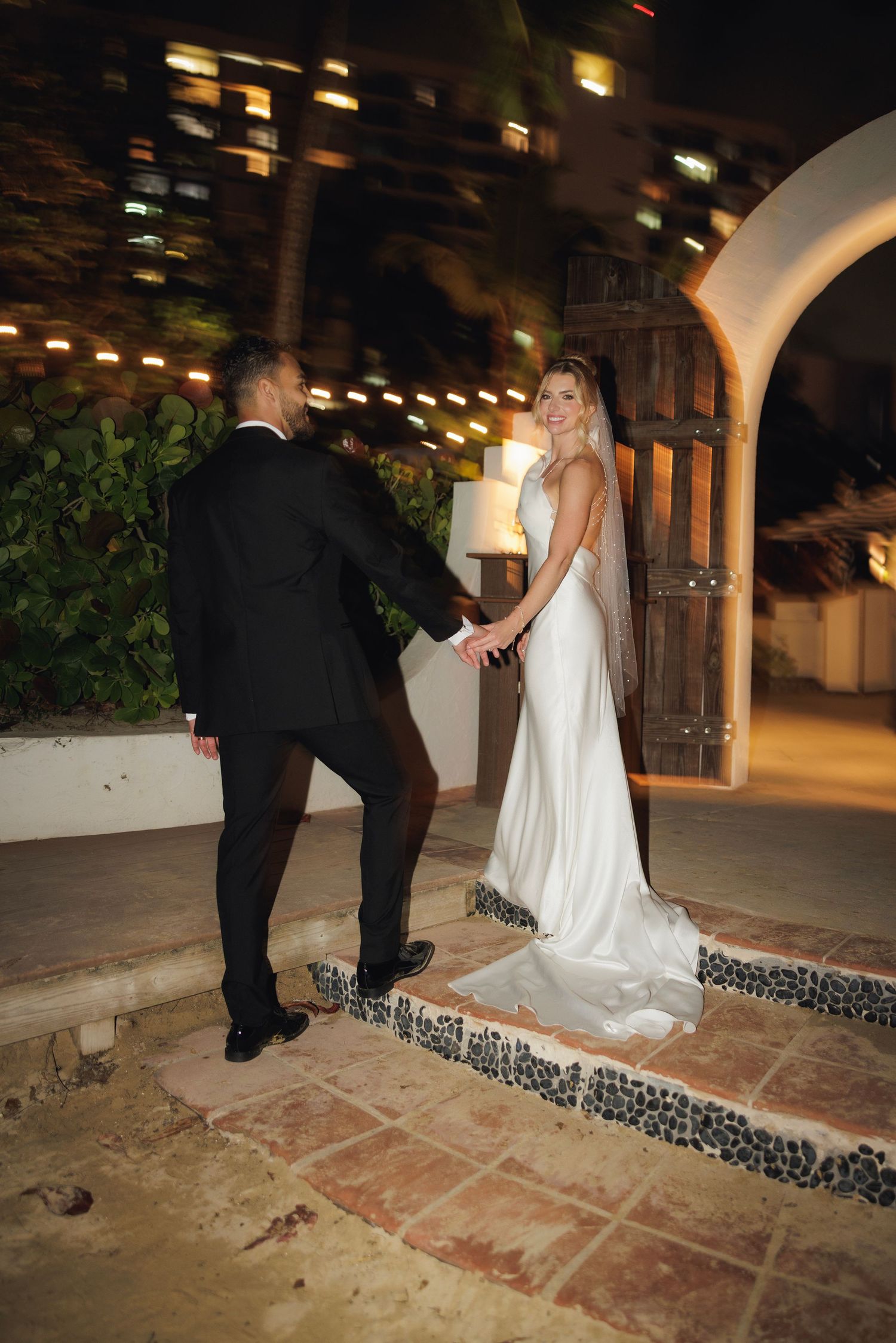 A couple shares an intimate moment at night under string lights on a tiled patio with arched doorways.