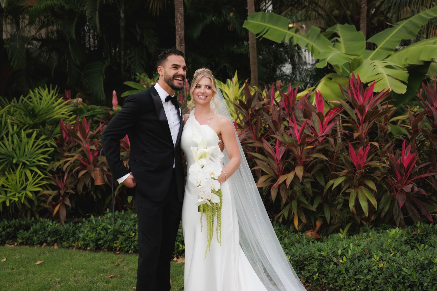 A newlywed couple stands together in a tropical garden setting with lush foliage and red plants in the background.