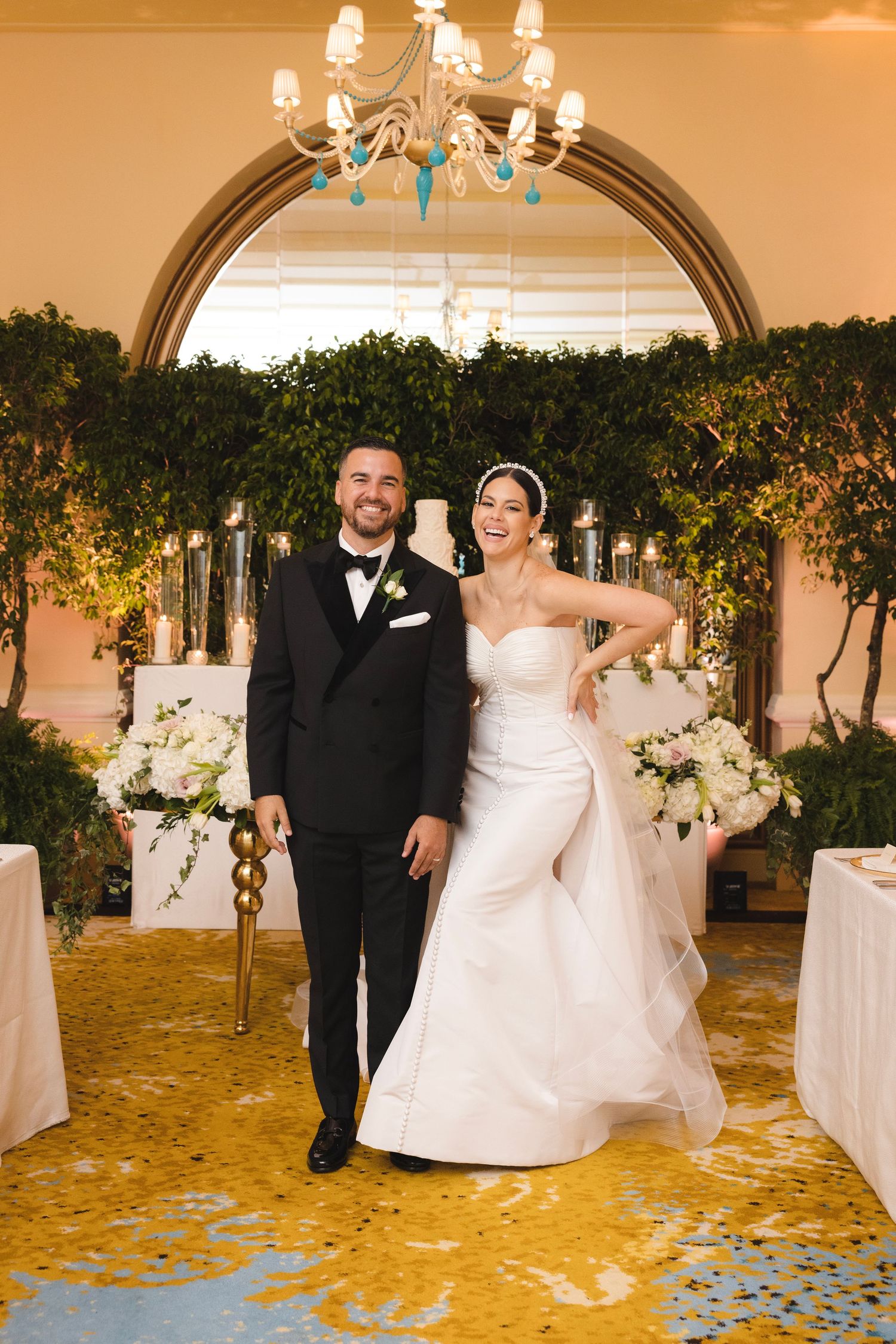 Wedding couple dances in an elegant ballroom with chandeliers and greenery decorations under warm lighting.