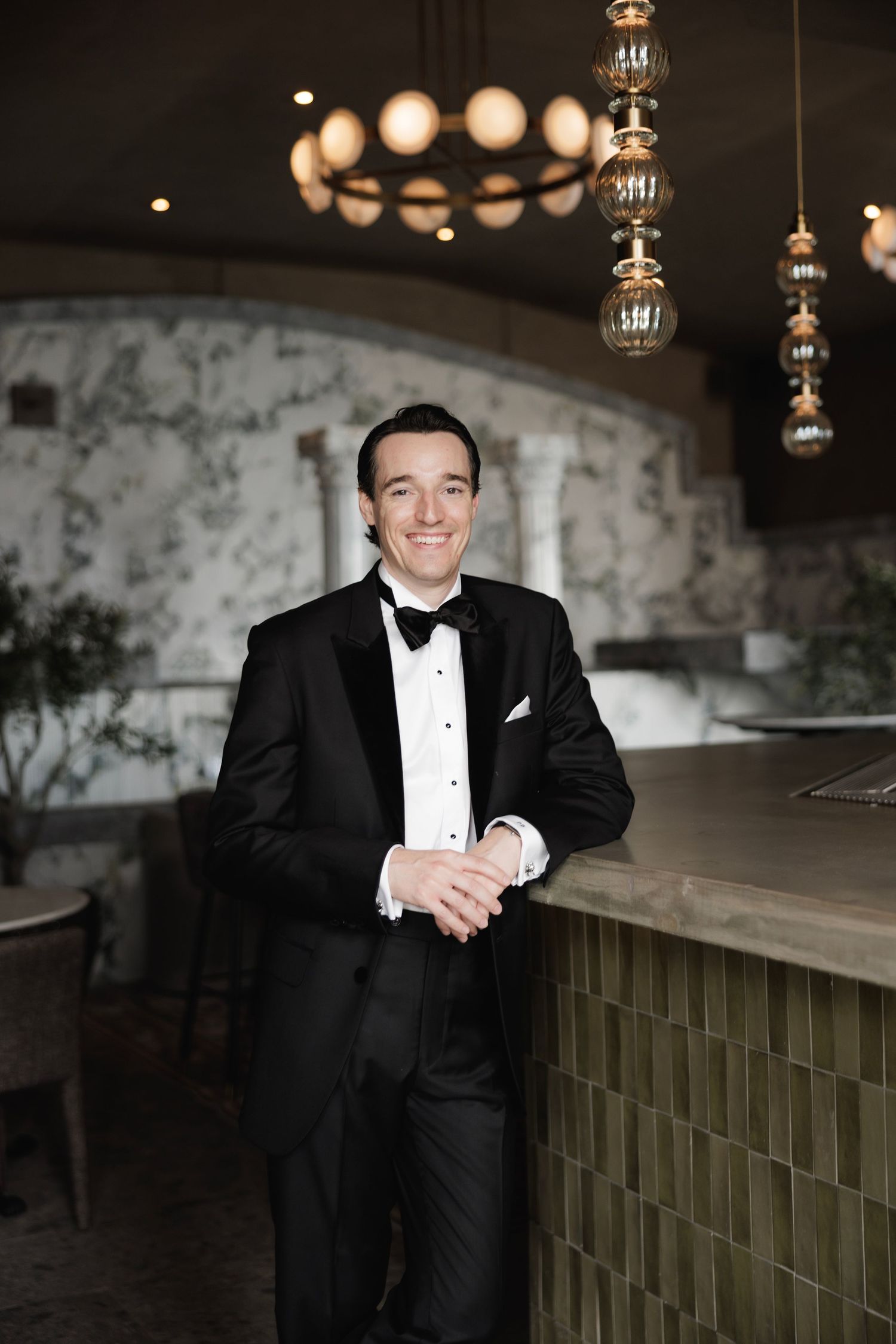A tuxedo-clad figure poses near illuminated pendant lights in an elegant venue setting.