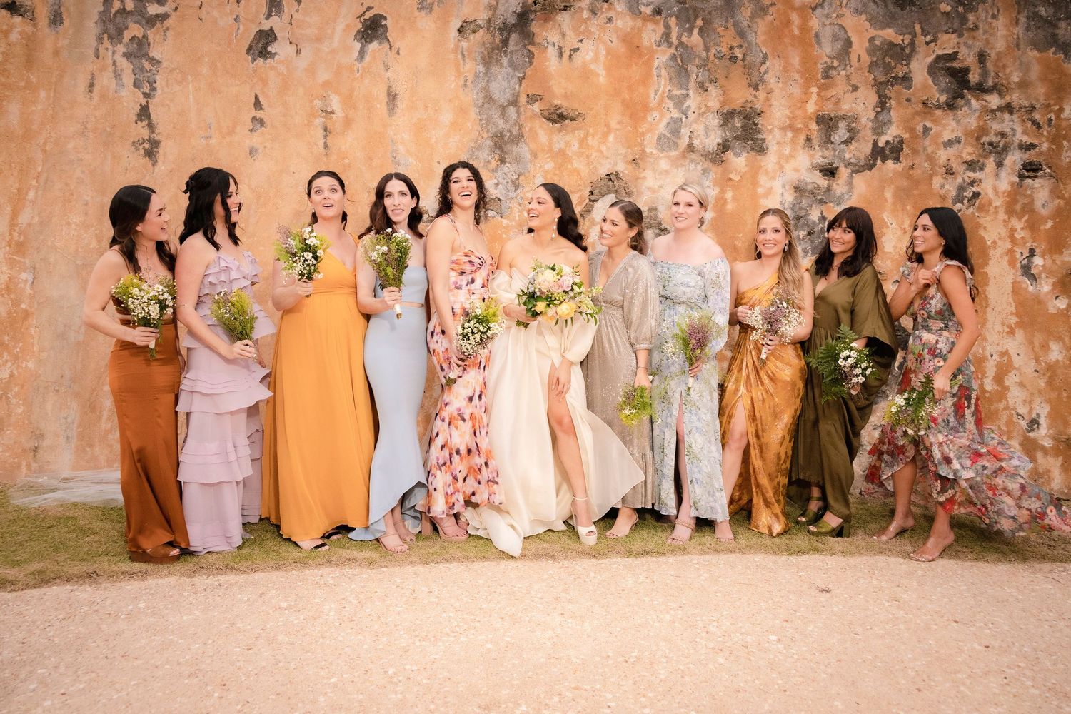Wedding party in pastel dresses poses against rustic stone wall backdrop.