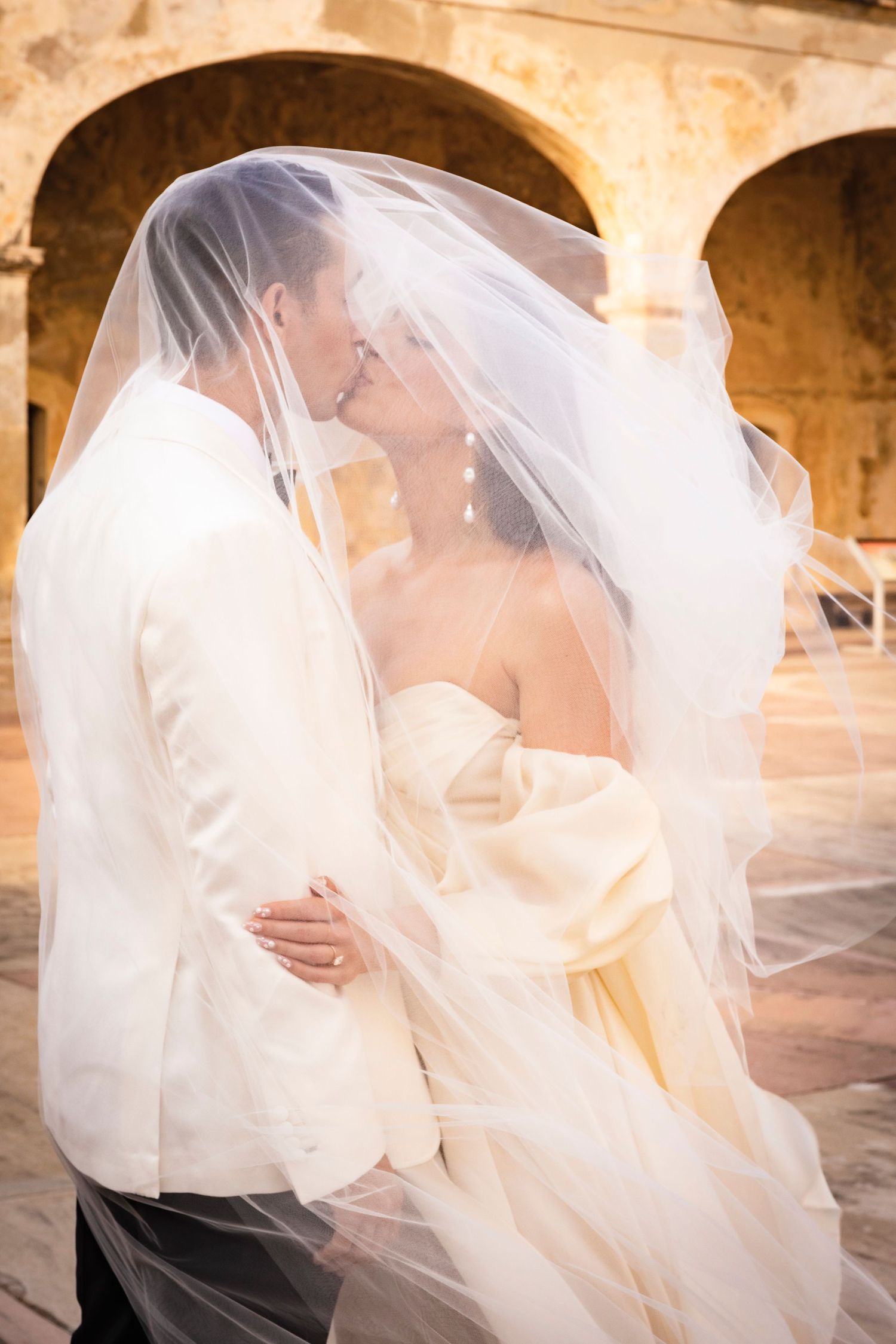 Romantic wedding moment captured through flowing veil in stone colonnade.