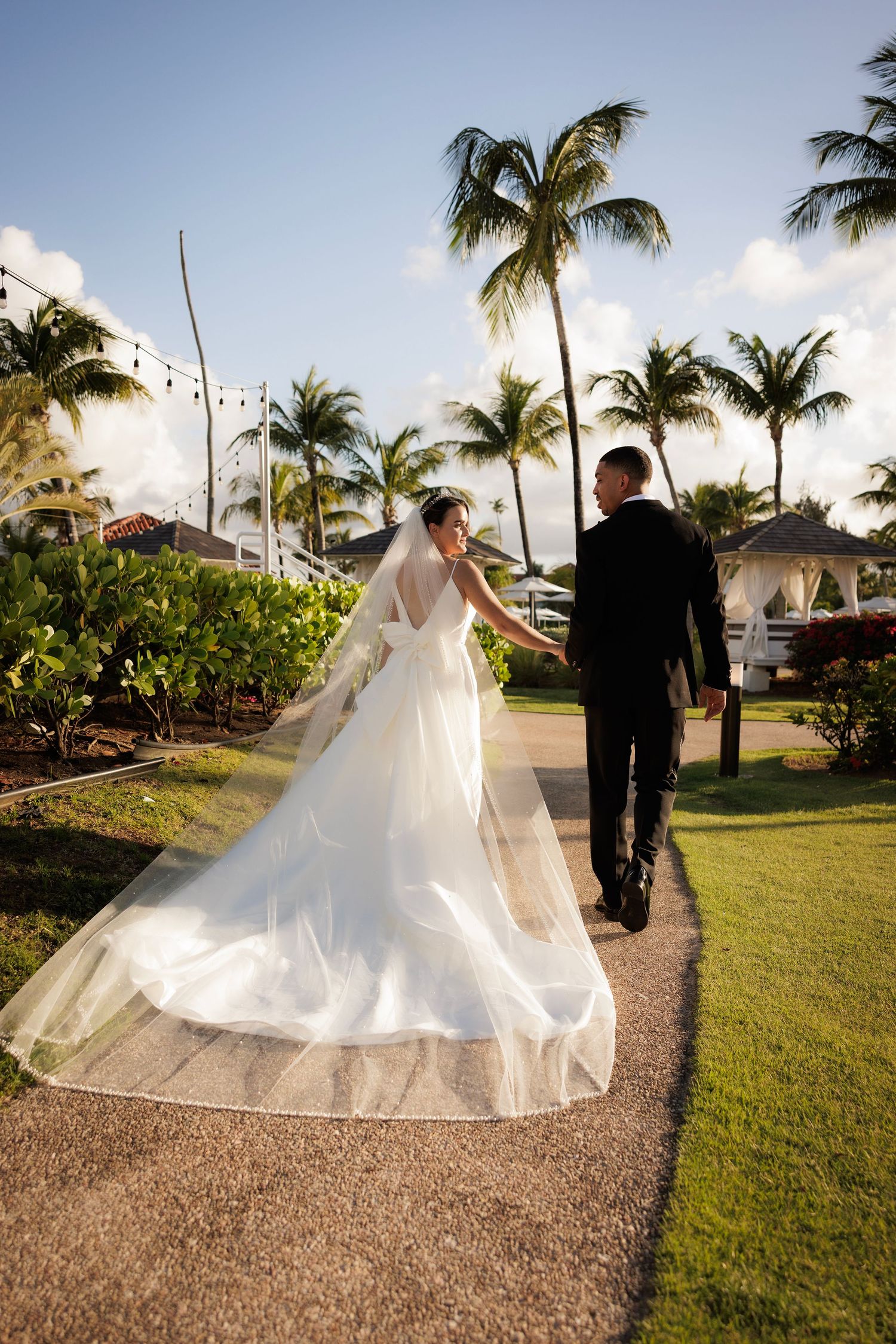Bride and groom walk along a palm-lined path at a tropical wedding venue during golden hour.