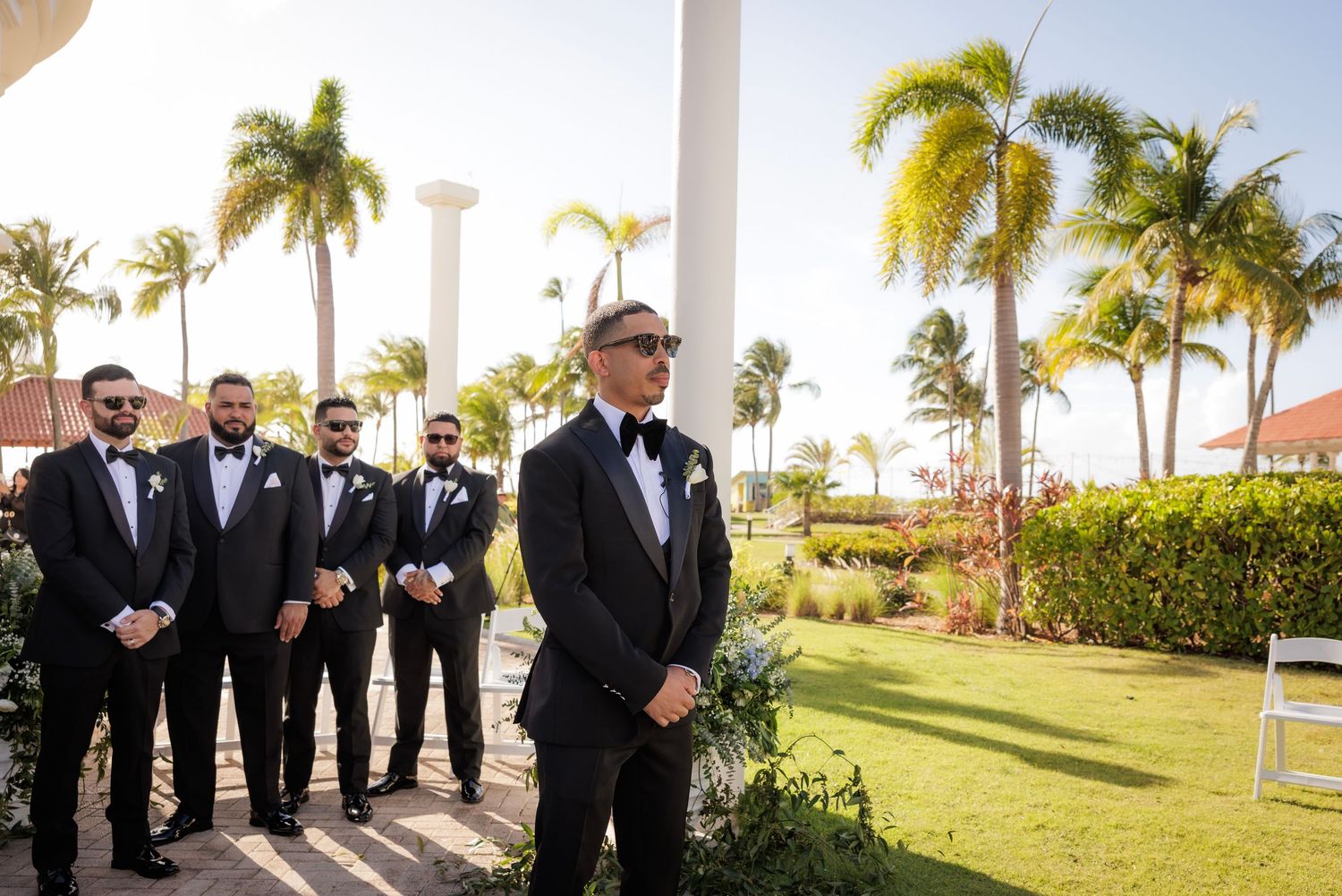 Groomsmen in formal black tuxedos pose together at an outdoor tropical venue with palm trees and white columns.