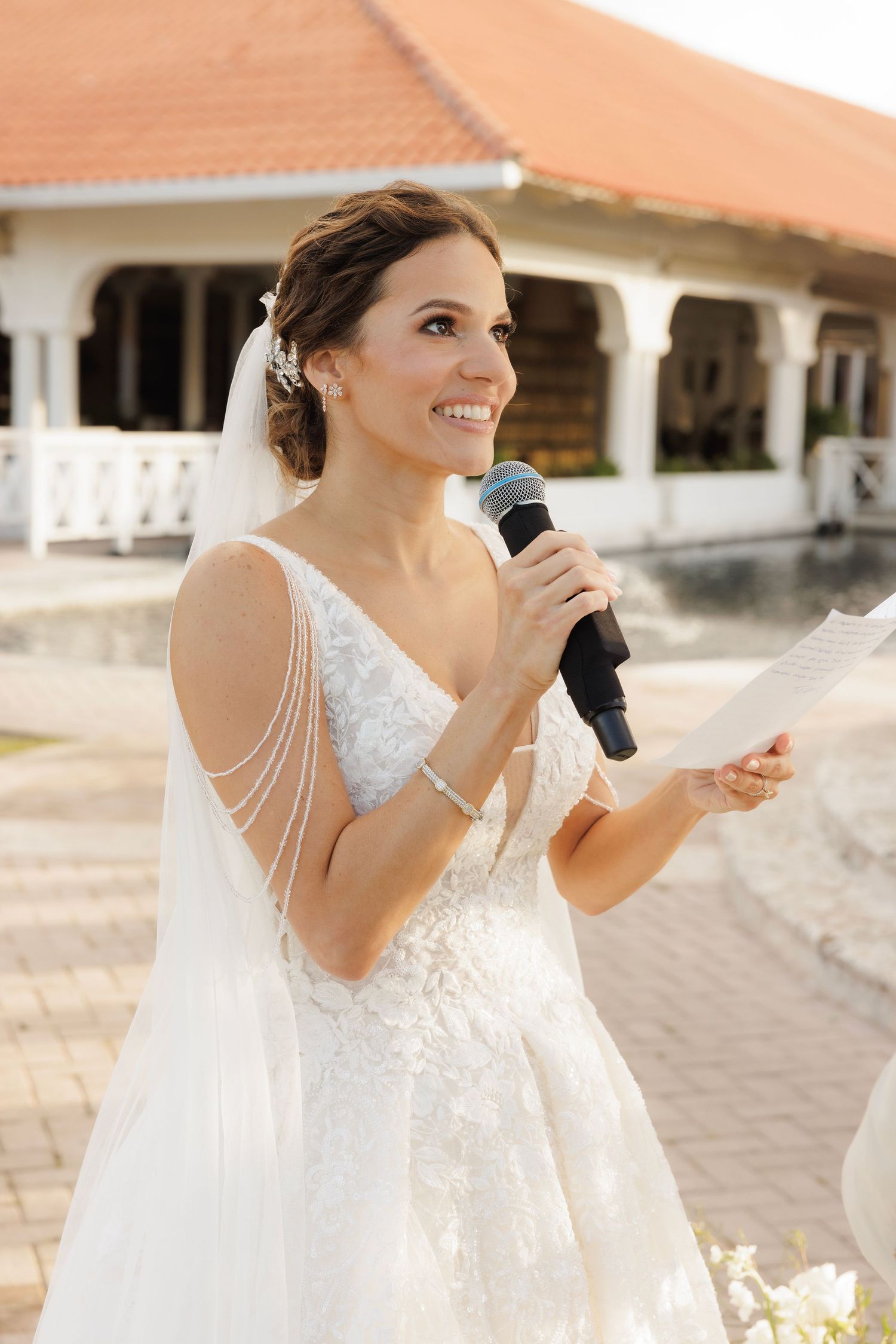 A bride in a flowing white lace wedding dress speaks into a microphone during an outdoor reception.
