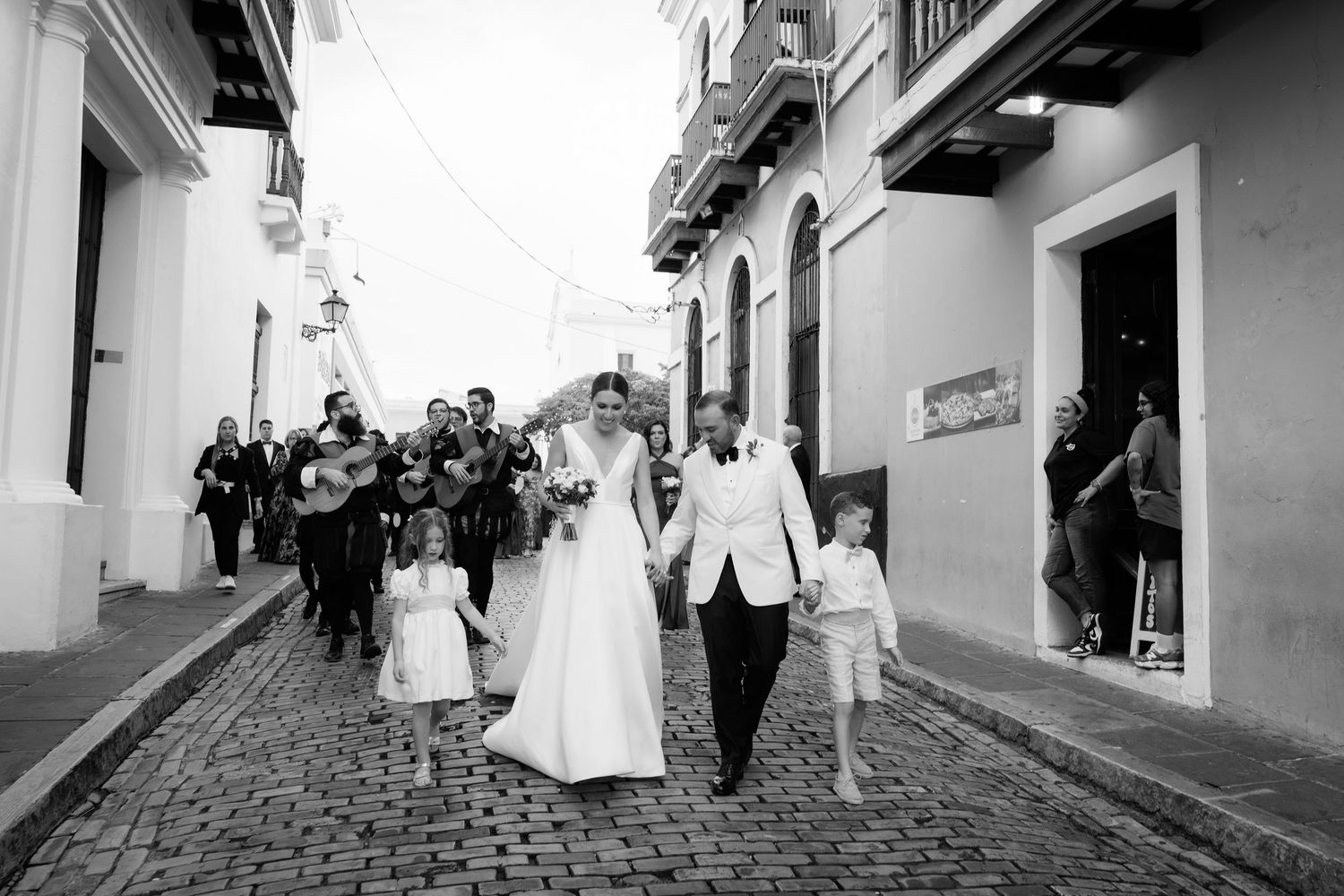 A wedding party walks down a narrow cobblestone street with white buildings.