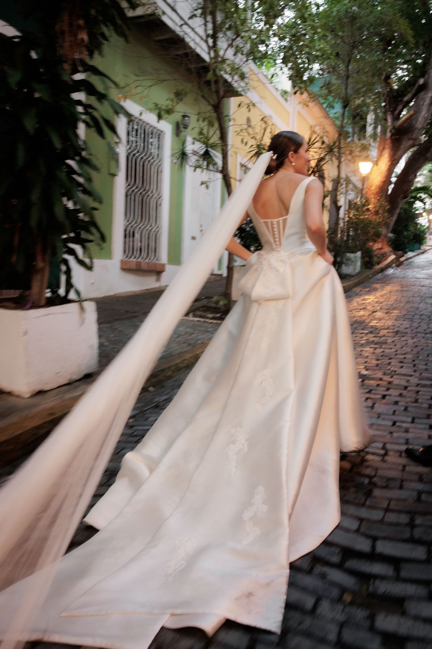 A flowing wedding dress train trails behind on a cobblestone street lined with pastel buildings.