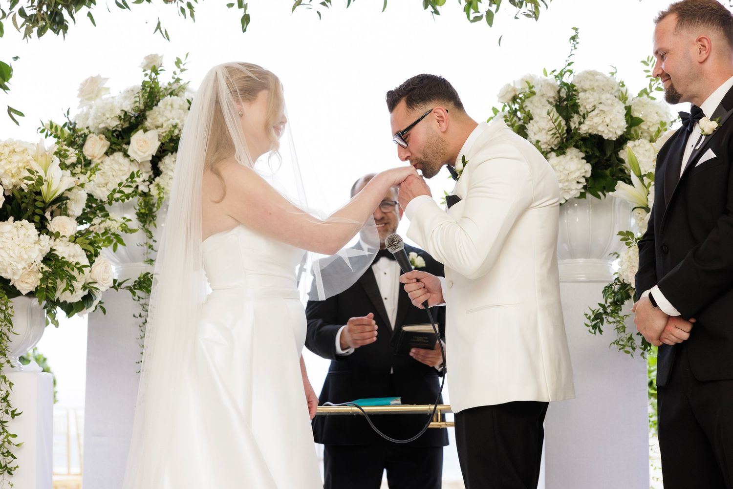 Wedding ceremony participants in formal attire standing among white floral arrangements.