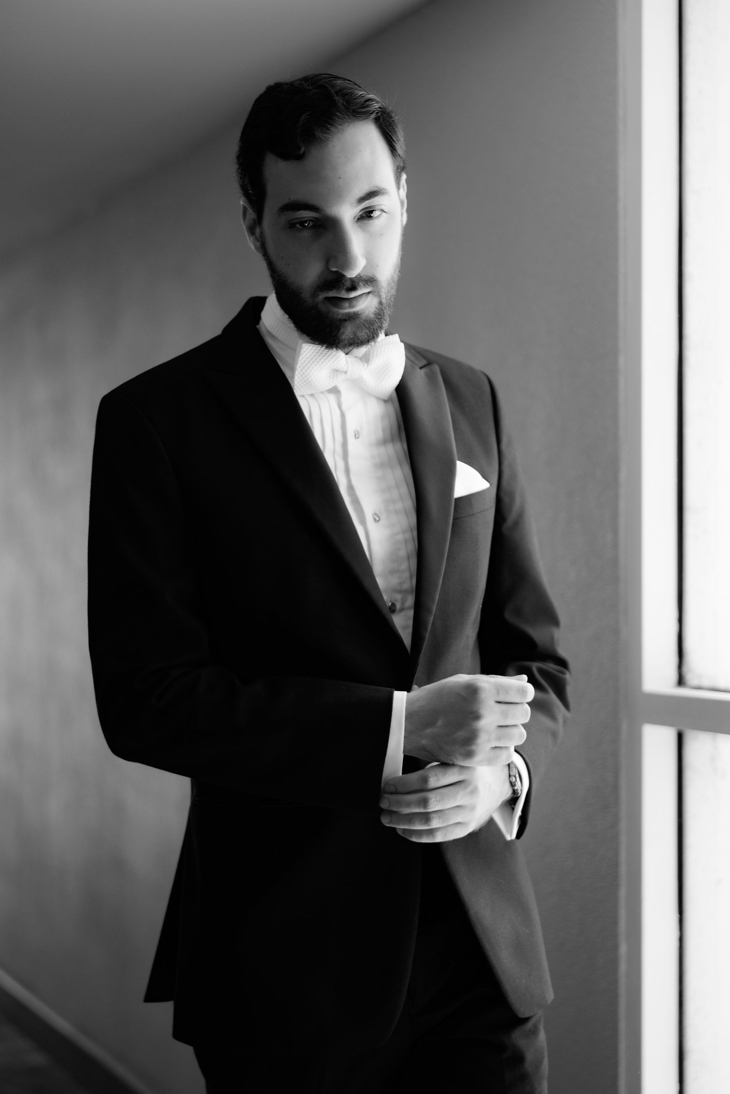 A series of black and white portraits of a groom in a formal tuxedo holding a drink near a window.