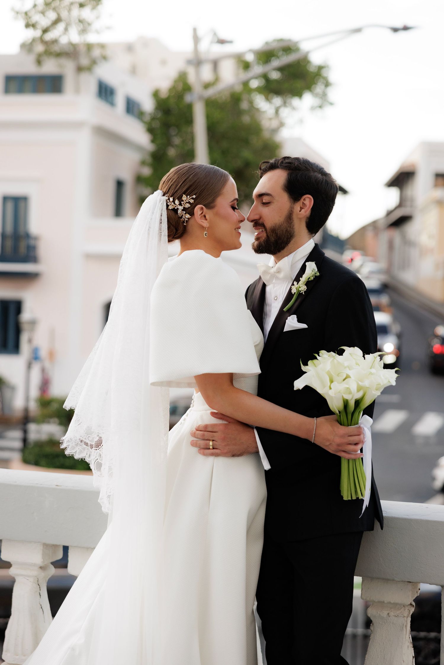 Newlywed couple share an intimate moment outdoors, bride in elegant white dress with cape holding calla lily bouquet.