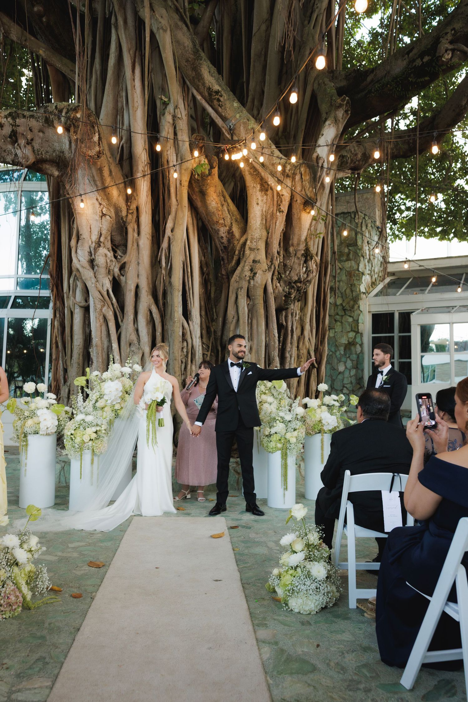 An outdoor wedding ceremony under a majestic banyan tree decorated with hanging string lights and white floral arrangements.
