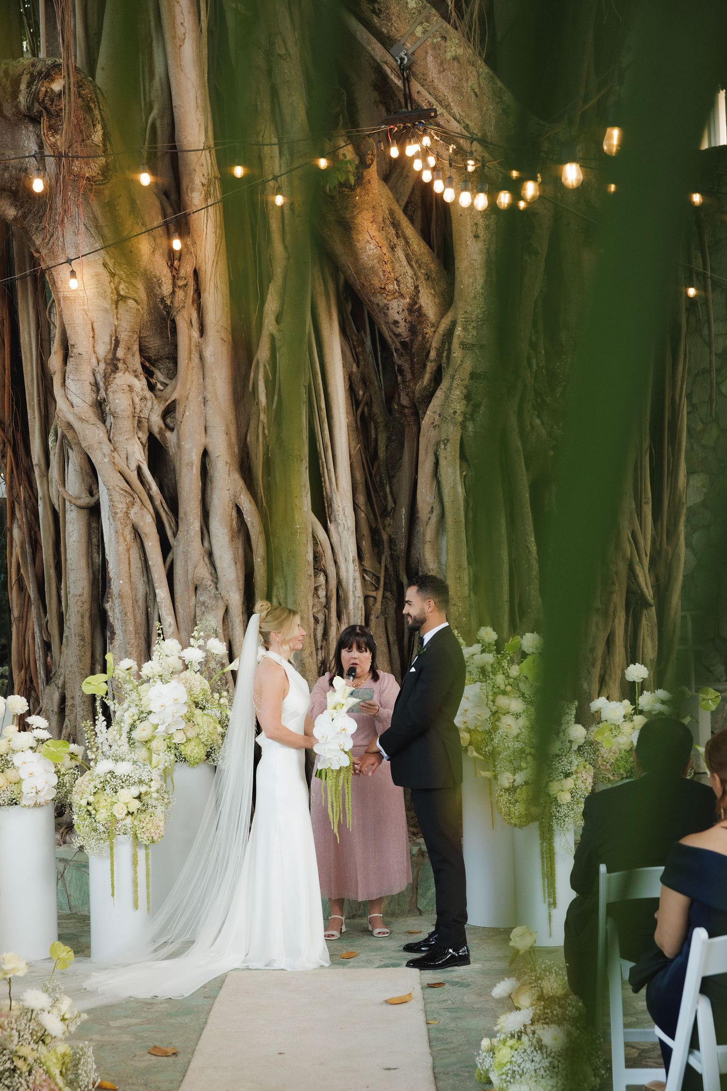 Wedding ceremony setup under a banyan tree with string lights and white floral arrangements creating a romantic atmosphere.