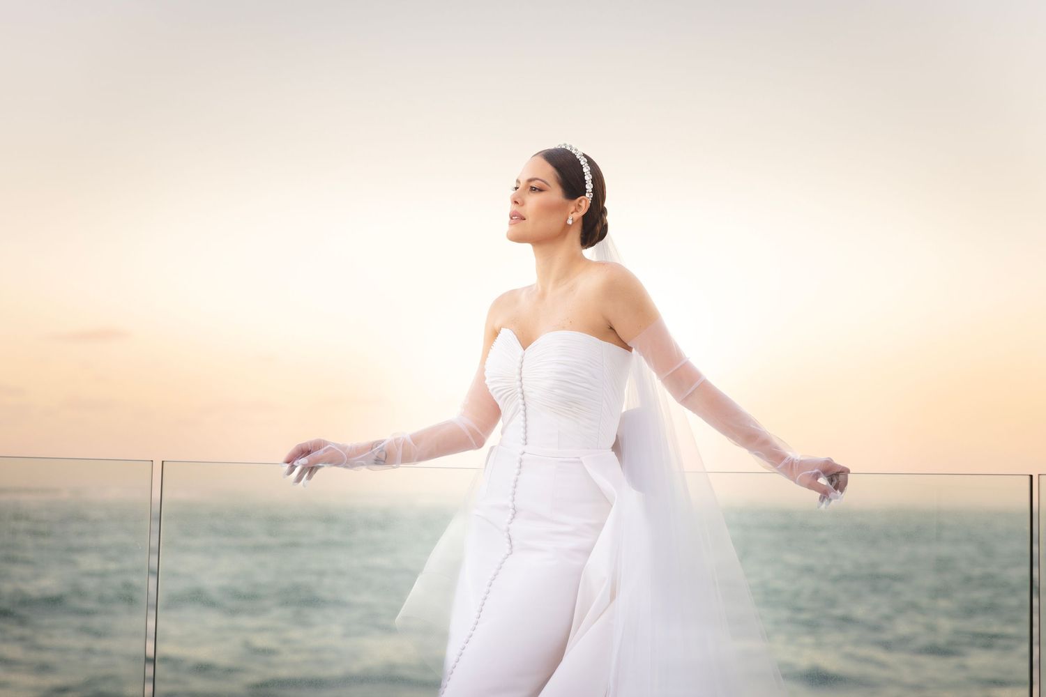 Woman in flowing white dress stands gracefully against a dreamy sunset backdrop at the beach.