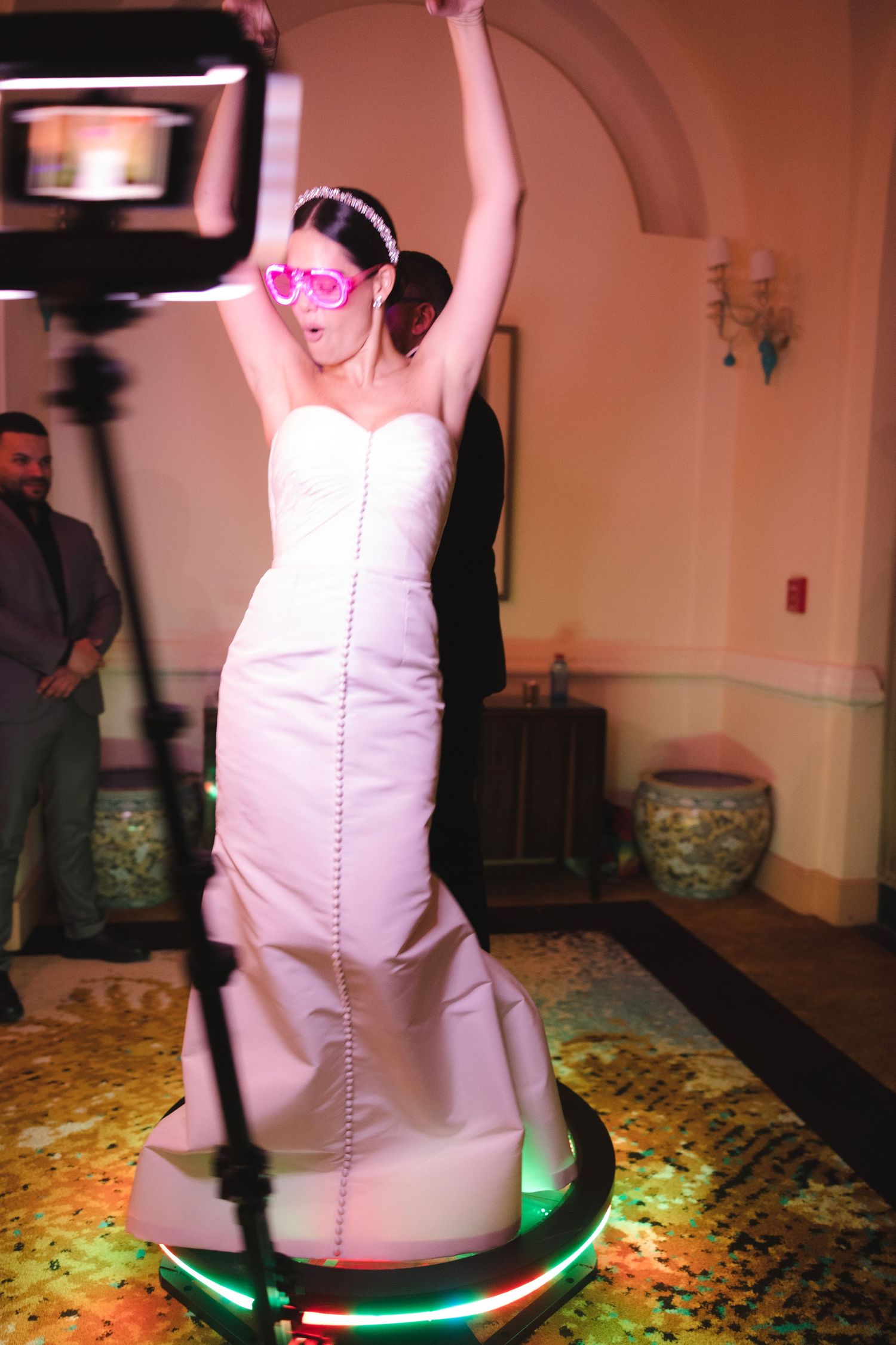 A person in a white wedding dress and pink sunglasses dances on a lit-up platform at a reception.