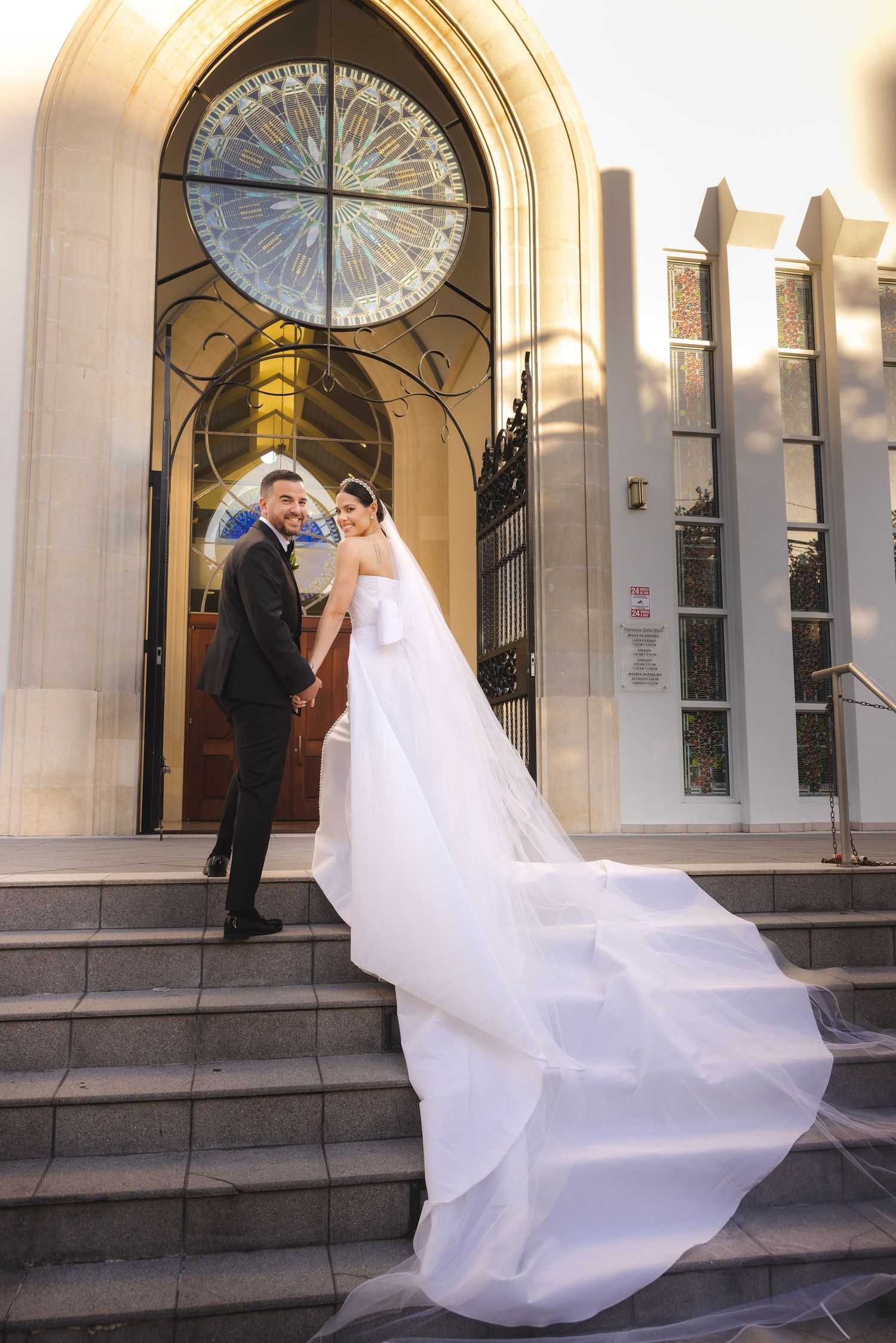 A bride's flowing white wedding dress dramatically billows in the breeze as she stands on stone church steps.