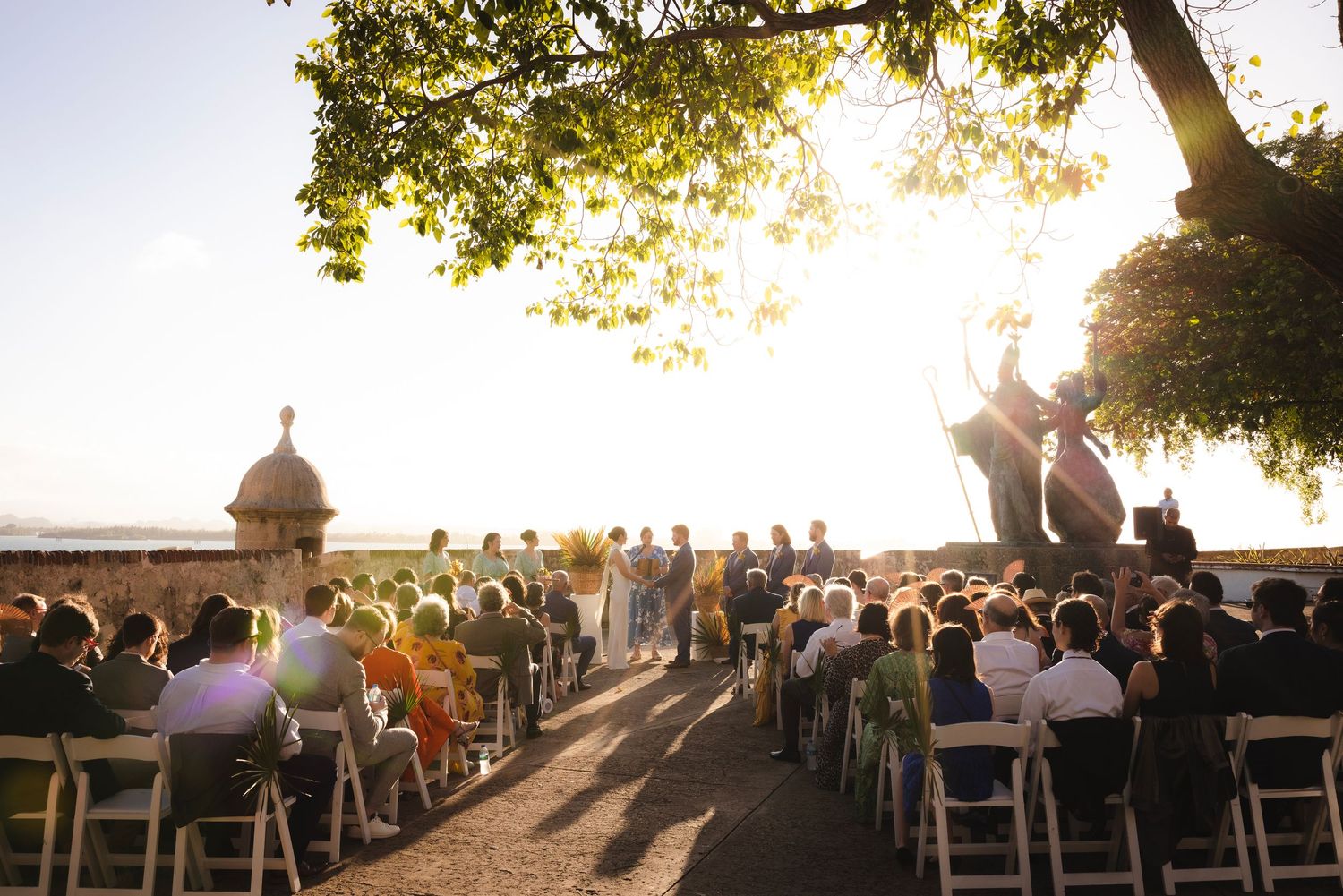 Wedding guests seated in wooden chairs at an outdoor ceremony during golden hour sunset.